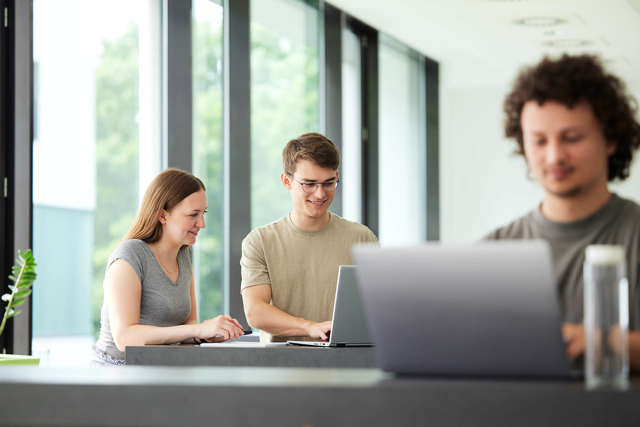 Im Fokus sind im Hintergrund zwei Studenten zu sehen die auf ihrem Laptop arbeiten, während rechts im Vordergrund unscharf ein Student ebenfalls mit seinem Laptop arbeitet