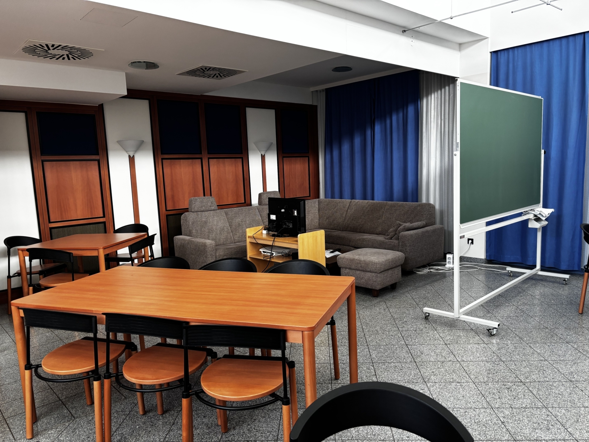 Ein Aufenthaltsraum mit einem Tisch im Vordergrund. Im Hintergrund eine Couch und eine Schreibtafel