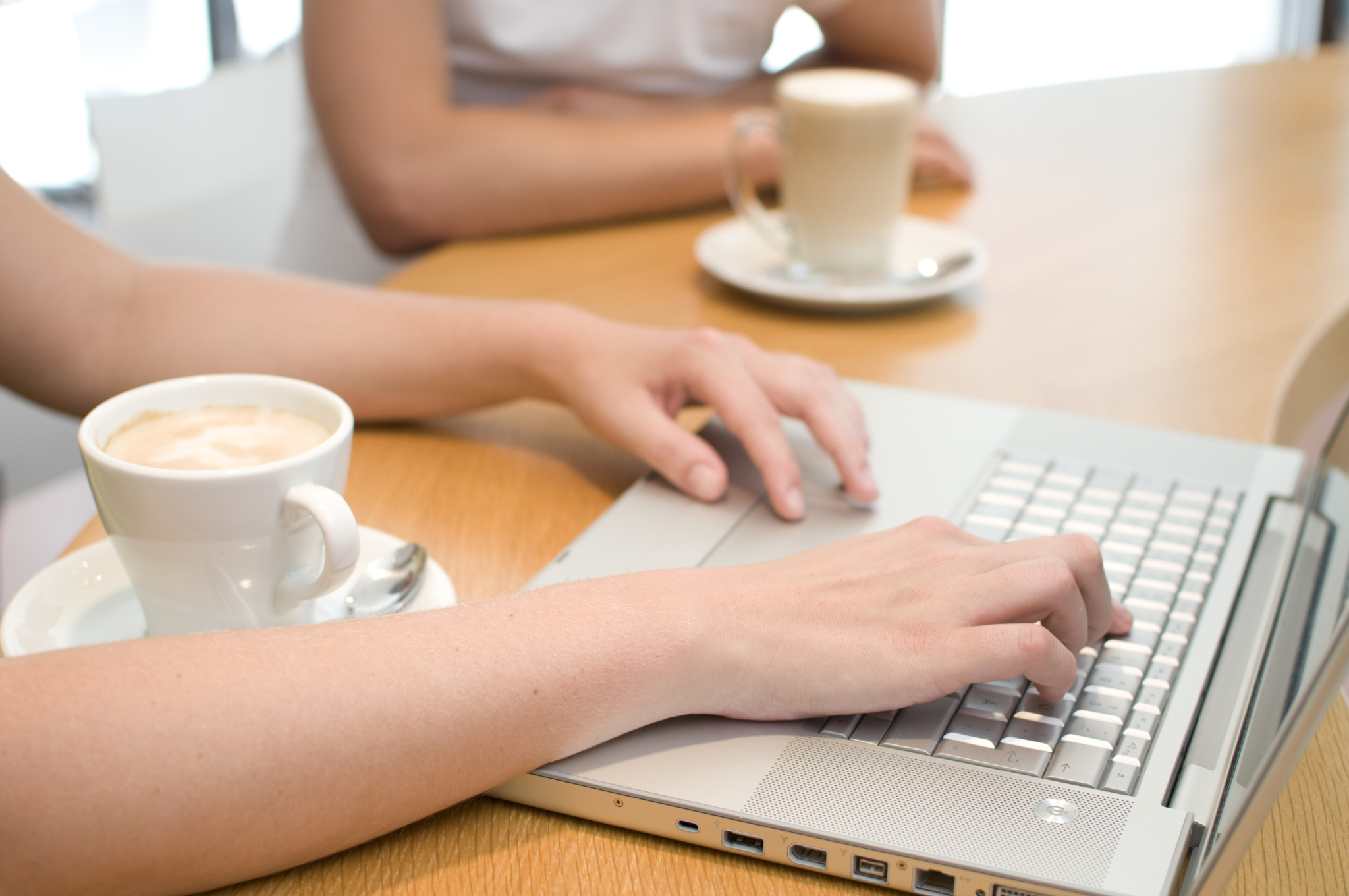 Im Fokus sind 2 Hände auf einer Laptop-Tastatur und davor steht eine Tasse Kaffee