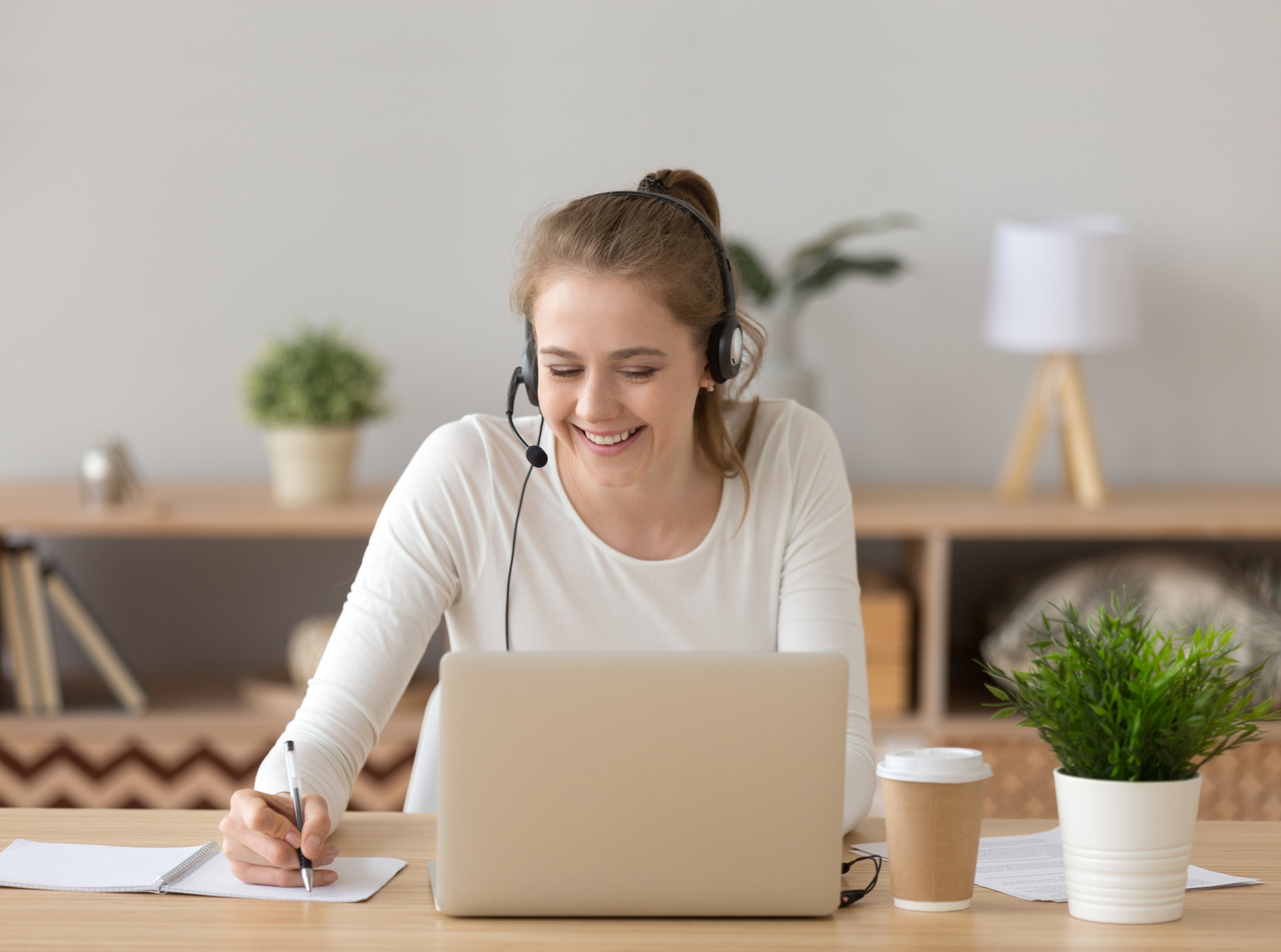 Eine Person sitzt mit einem Headset vor dem Laptop und notiert sich etwas auf einem danebenliegenden Notizblock