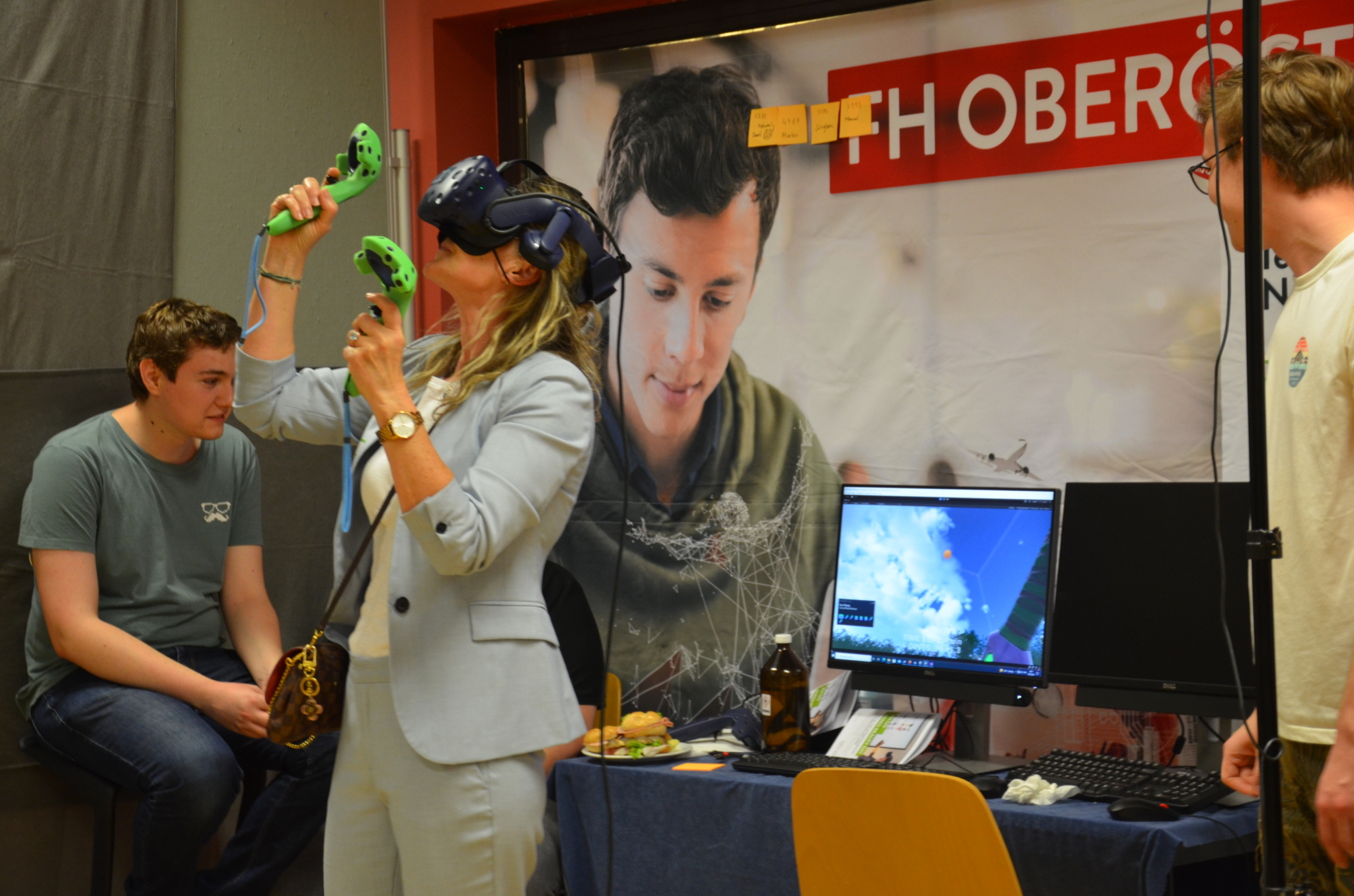 Eine Person steht neben einem Computer, hat eine VR-Brille auf, zwei Joysticks in der Hand und spielt in einer virtuellen Welt.