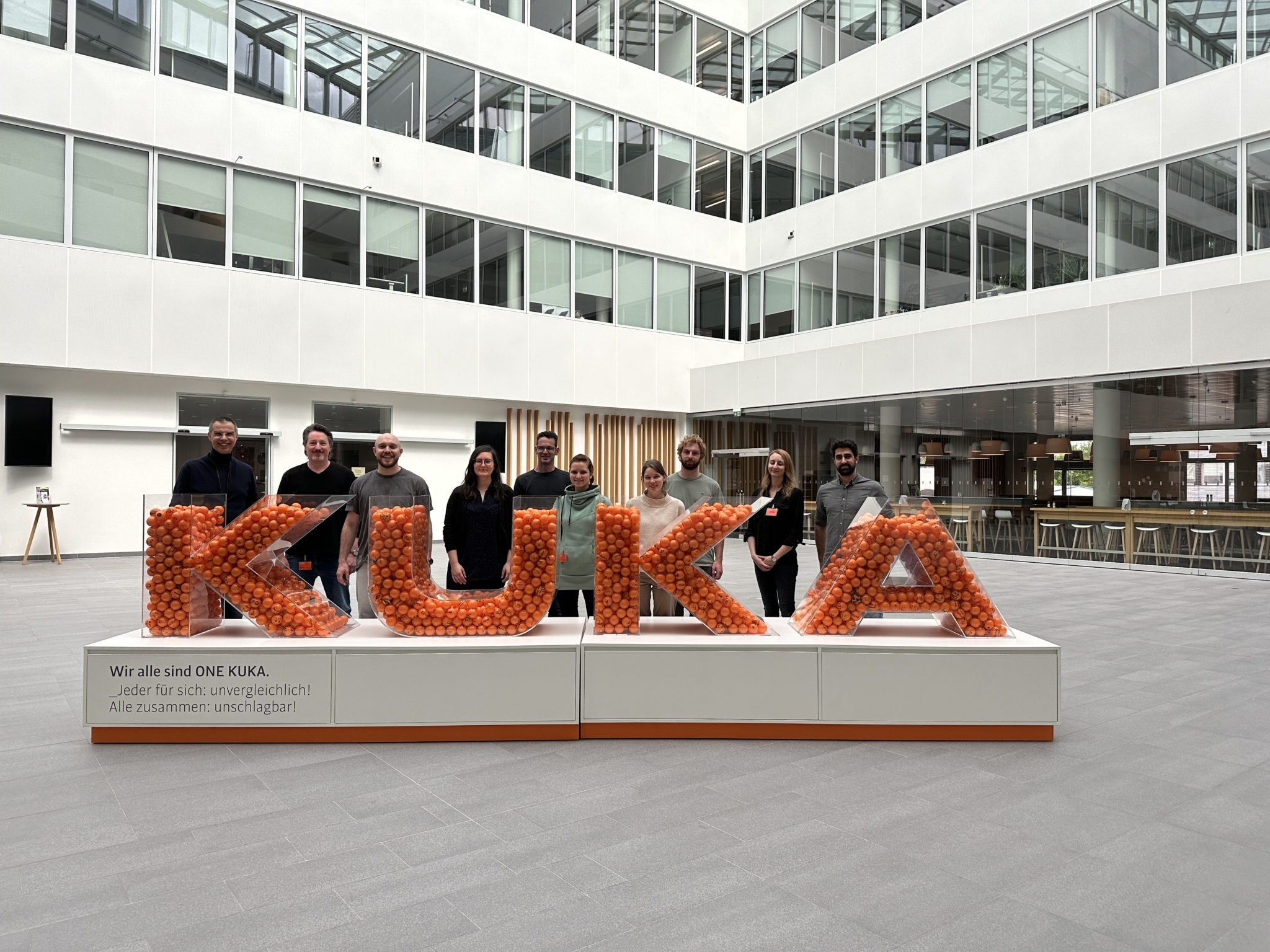KUKA Schriftzug aus Plexiglas gefüllt mit orangen Bällen und dahinter stehen ein Gruppe Menschen mit Blick in die Kamera