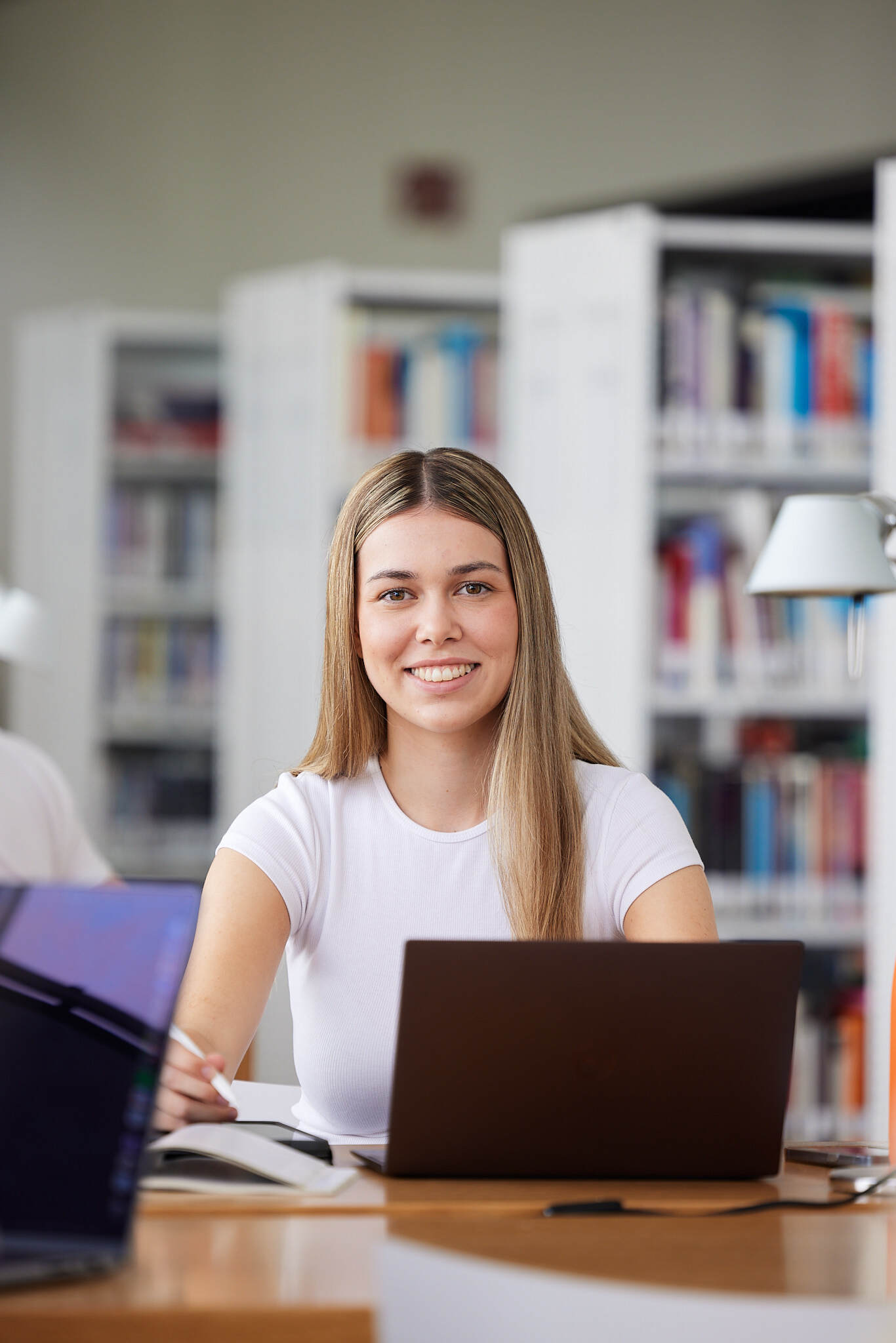 Eine Studentin sitzt in der Bibliothek vor einem geöffneten Laptop und lächelt in die Kamera