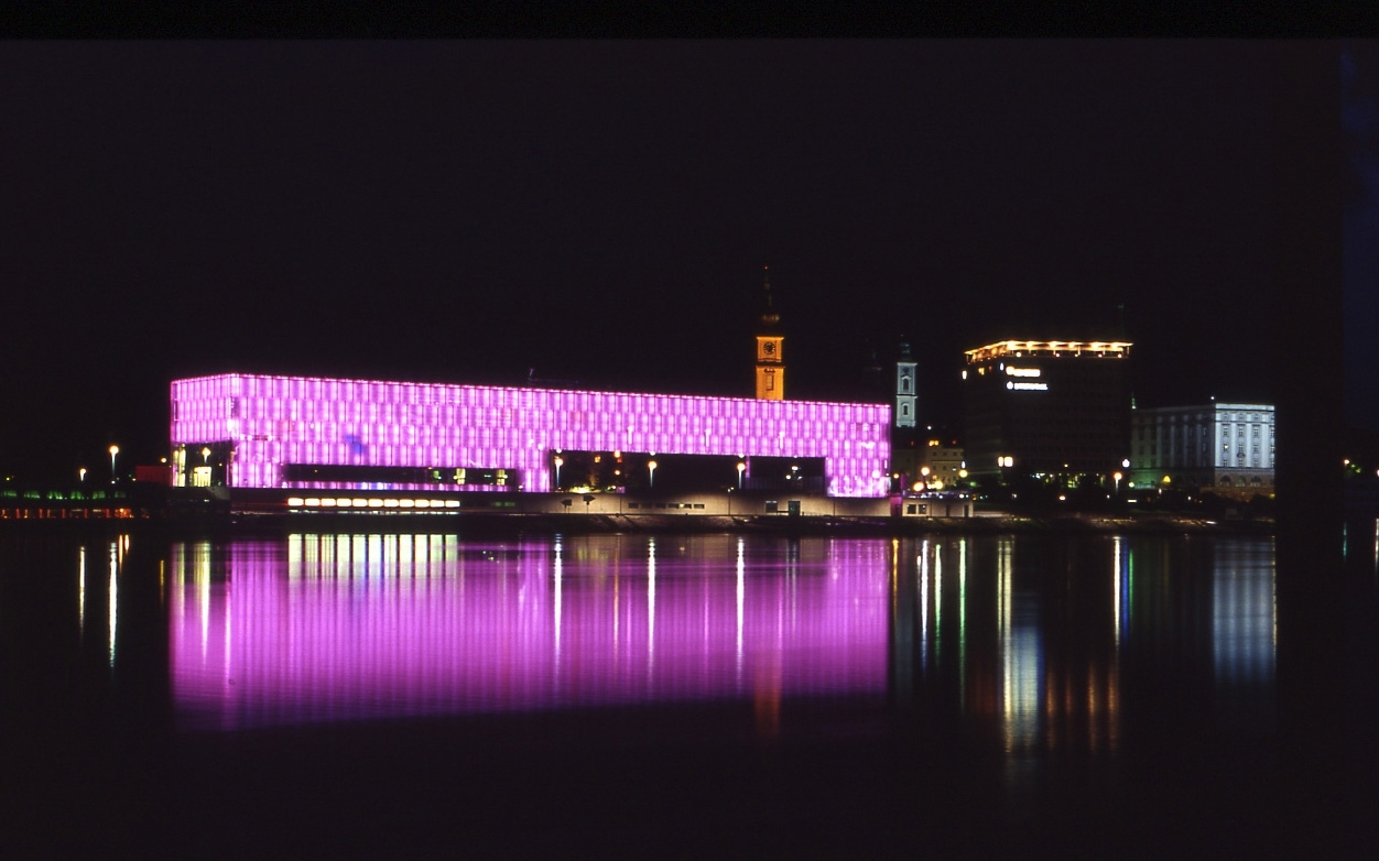 Das Lentos wird bei Nacht in Lila Farben beleuchtet und von der anderen Uferseite der Donau aus gezeigt