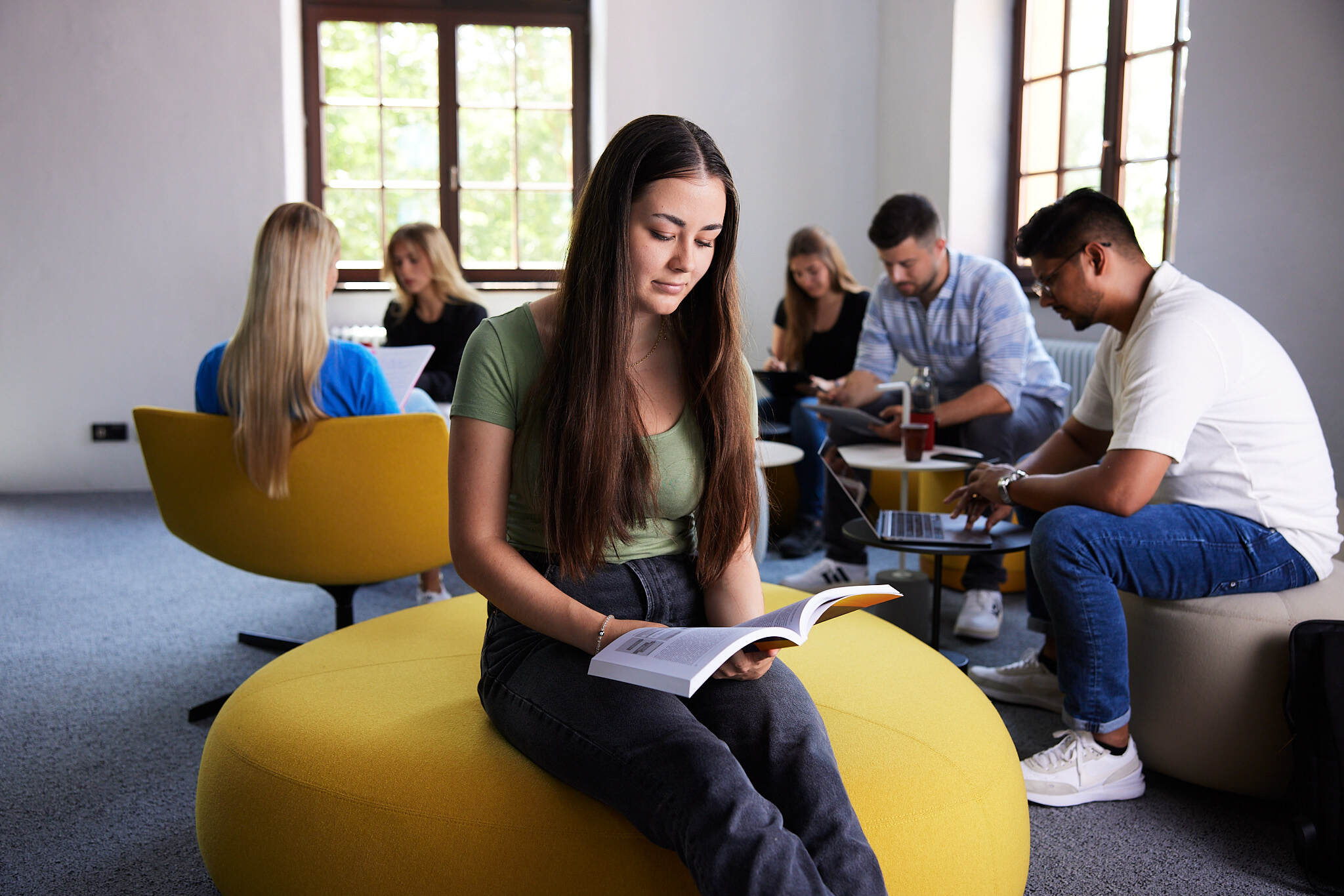 Im Fokus ist eine junge Studentin die auf einem Hocker in einem Aufenthaltsraum am Campus Steyr sitzt, während die anderen Studenten im Hintergrund unscharf sind.