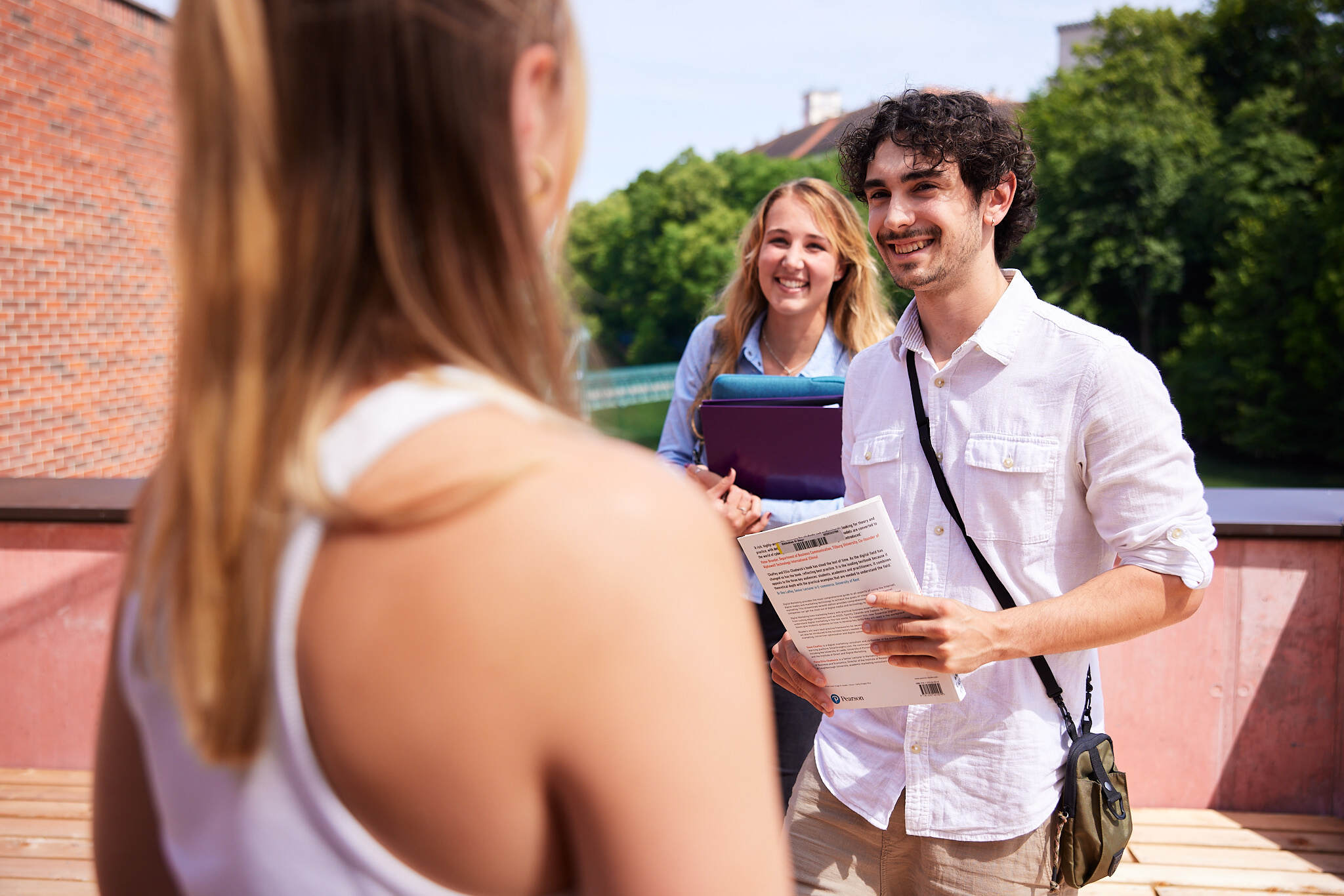 zwei junge Studenten gehen auf eine unscharfe Studentin im Vordergrund zu, auf einer Terrasse vor dem Fluss Steyr mit Büchern und Laptops in ihren Händen.