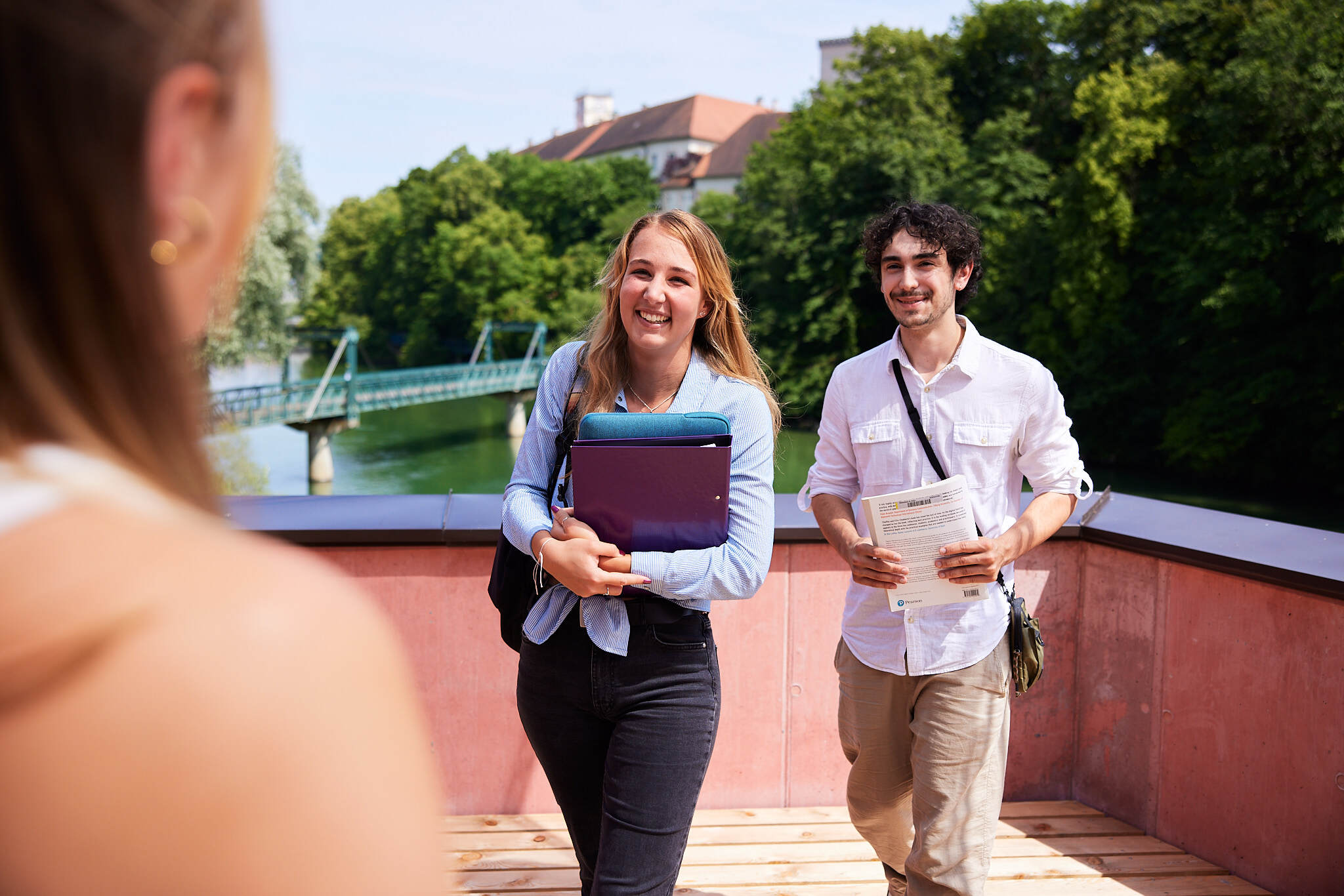 zwei junge Studenten gehen auf eine unscharfe Studentin im Vordergrund zu, auf einer Terrasse vor dem Fluss Steyr mit Büchern und Laptops in ihren Händen.