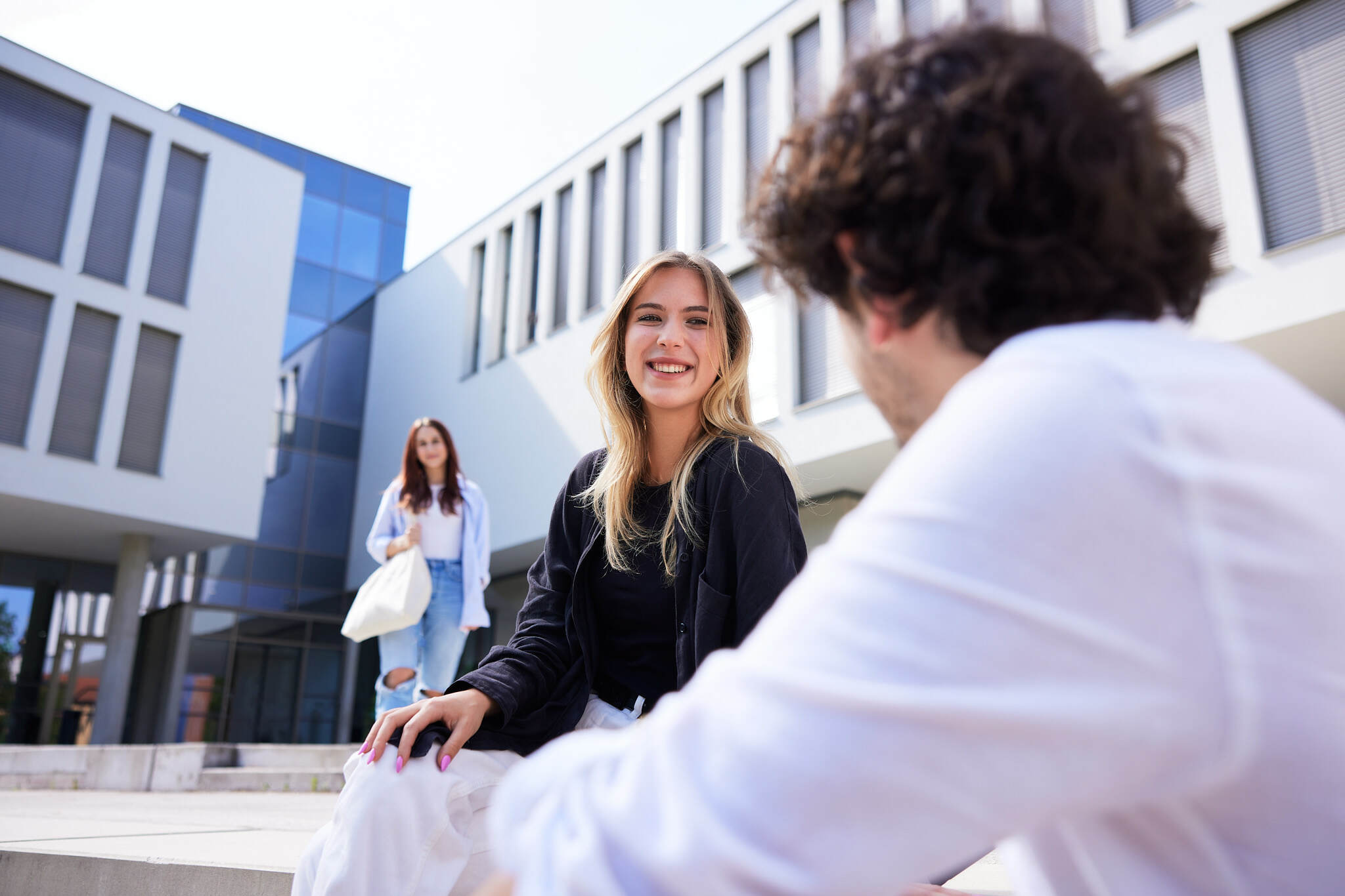 eine junge Studentin sitzt vor einem Campusgebäude der FH Steyr, während sie sich mit einem Studenten, der unscharf im Vordergrund ist, unterhält.