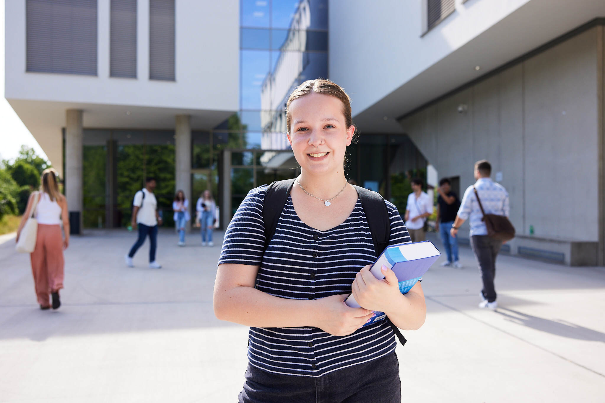 Eine junge Studentin steht mit einem Buch in der Hand vor einem Campusgebäude der FH Steyr.