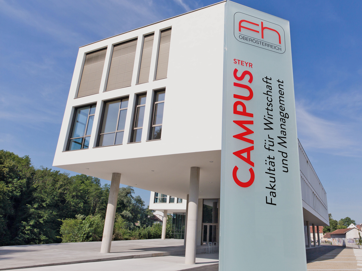 Außenaufnahme des FH3-Gebäudes der FH OÖ in Steyr, im Vordergrund eine Beschilderung mit der Aufschrift "Campus Steyr – Fakultät für Wirtschaft und Management"