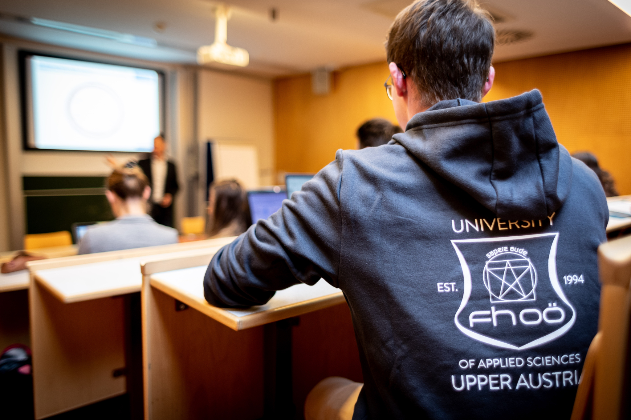 Ein Student sitzt in einem Hörsaal, von hinten fotografiert, mit einem Pullover der Fachhochschule Oberösterreich