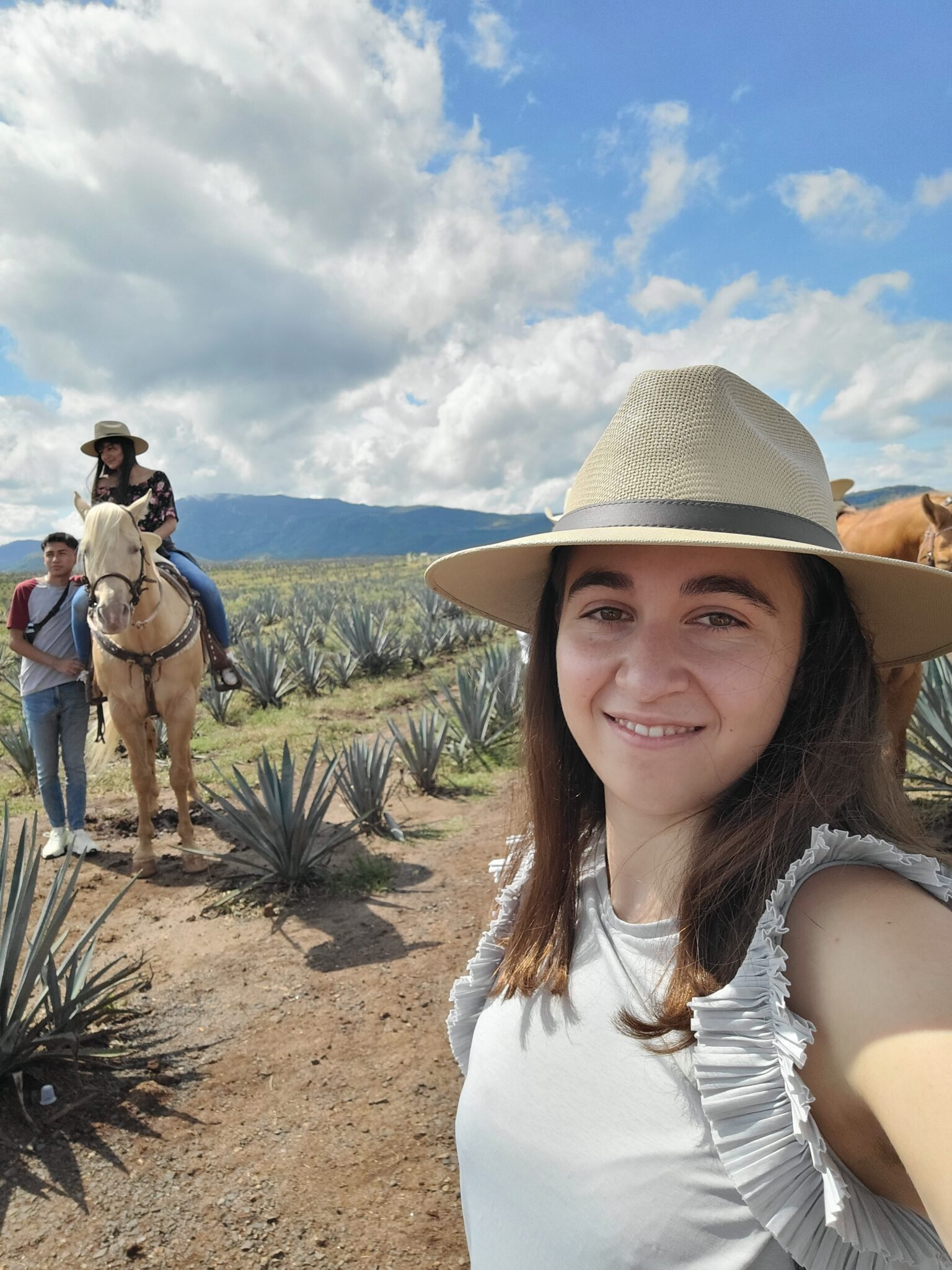 A young girl takes a selfie wearing a sun hat. Behind her, a boy stands next to a horse and another girl is sitting on a horse