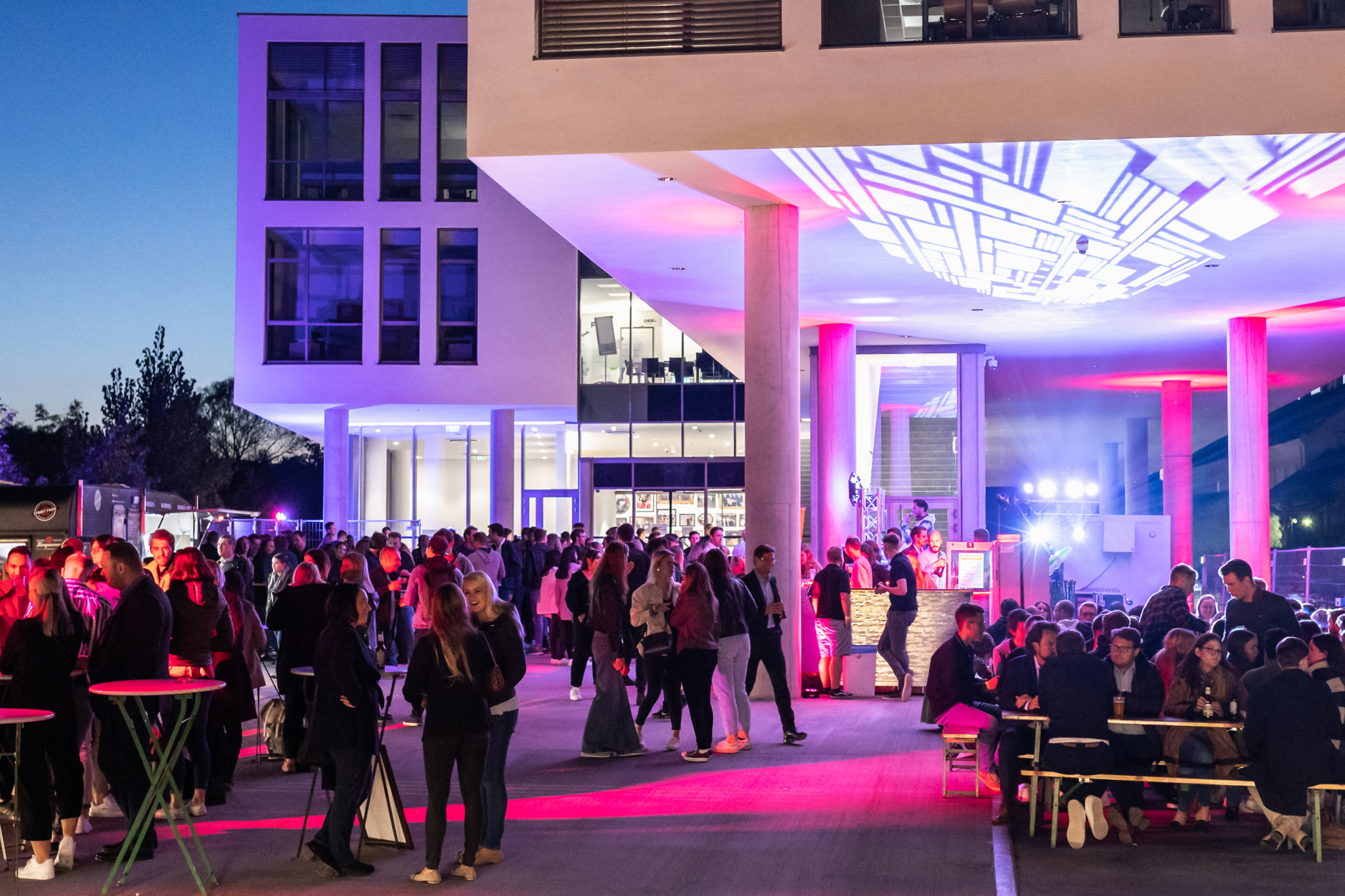 Foto von der Afterparty der Karrieremesse, auf dem viele Studierende auf Bierbänken sitzen und an Stehtischen stehen, umgeben von Partylichtern