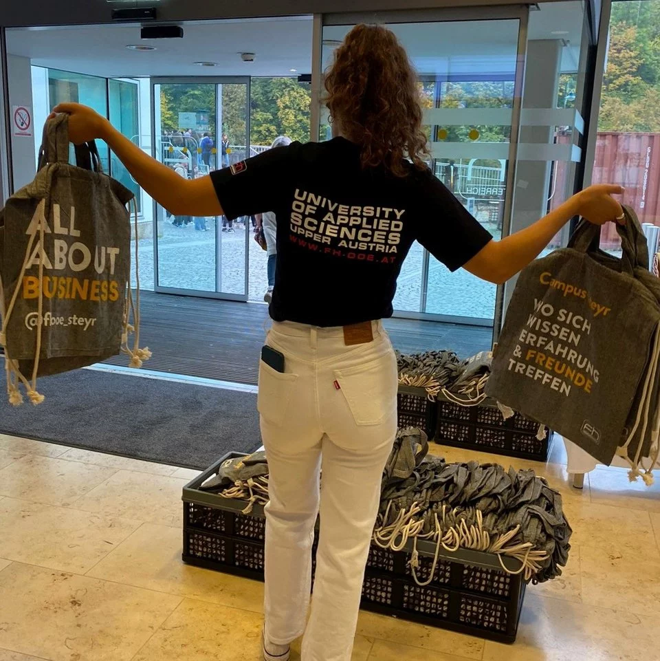 Foto einer jungen Frau von hinten, mit FH-Shirt und mehreren Goodie Bags in der Hand