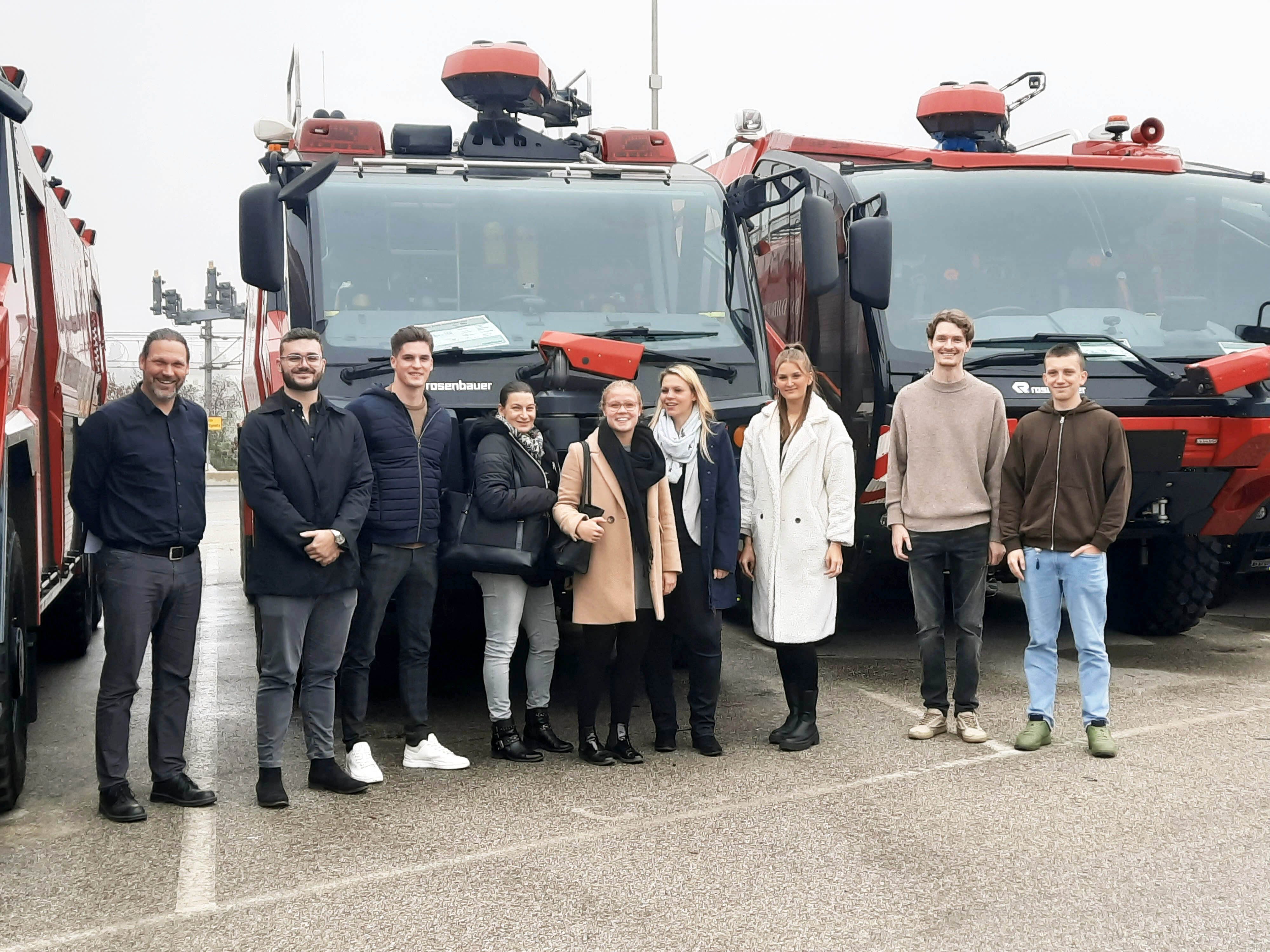 Auf dem Foto stehen neun Männer und Frauen vor Feuerwehrautos der Firma Rosenbauer und lächeln für ein Gruppenfoto.