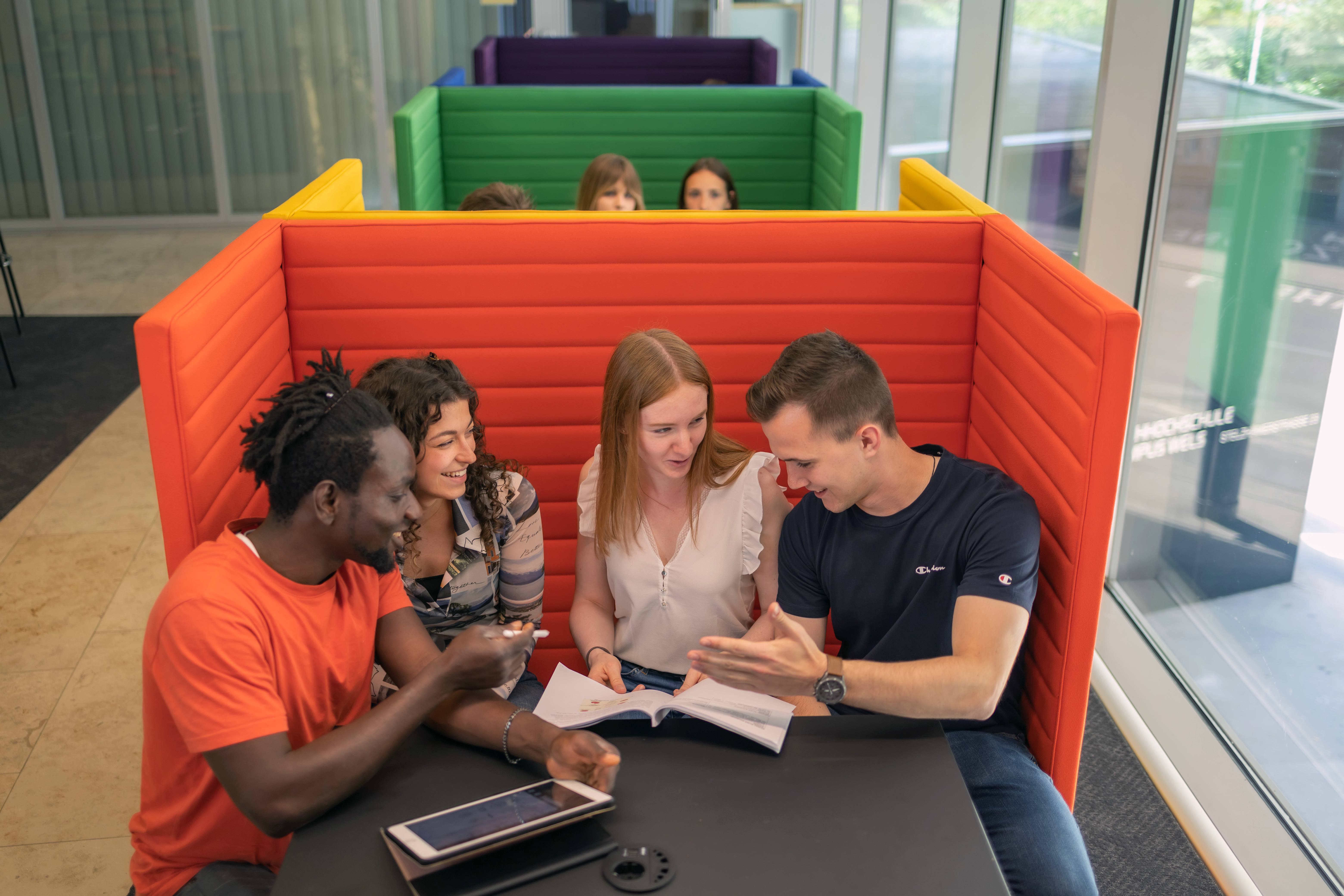 4 Studenten auf einer der Regenbogen-Bänke im Indoor-Bereich der Studylounge der FH Wels