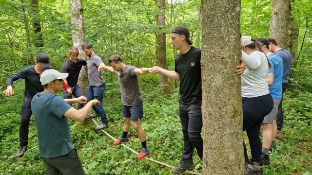 Acht Personen balancieren auf einer Slack-Line zwischen mehreren Bäumen während eine weitere Person Anweisungen gibt.