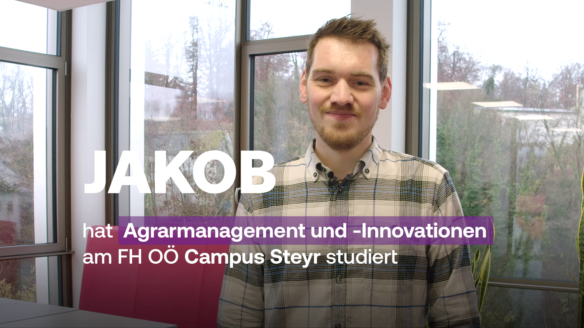 Ein junger Mann namens Jakob steht vor großen Fenstern und lächelt in die Kamera. Im Bild ist der Text eingeblendet: „JAKOB hat Agrarmanagement und -Innovationen am FH OÖ Campus Steyr studiert“. Im Hintergrund sind Bäume und Gebäude außerhalb der Fenster zu sehen.