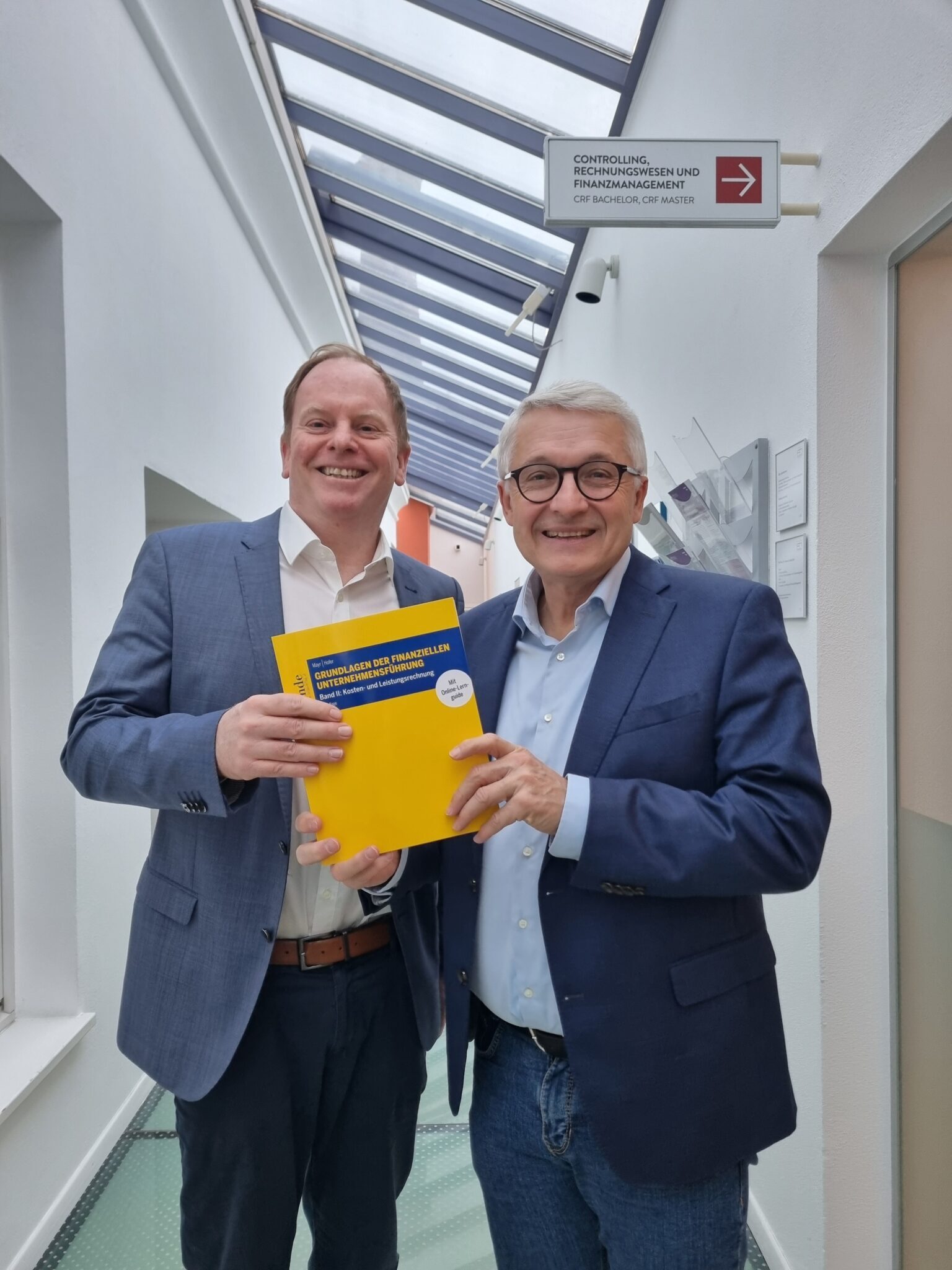 Albert Mayr und Peter Hofer im FH1 Gebäude halten die neue Auflage des Lehrbuchs "Kosten- und Leistungsabrechnung" in der Hand und schauen in die Kamera