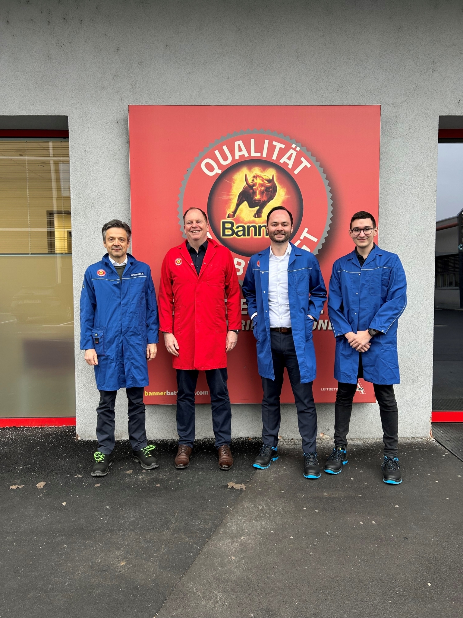 Vier Männer in blauer und roter Arbeitskleidung stehen nebeneinander vor einer Wand mit einem großen roten Schild mit dem Banner-Logo und dem Schriftzug ‚Qualität‘. Sie lächeln in die Kamera.