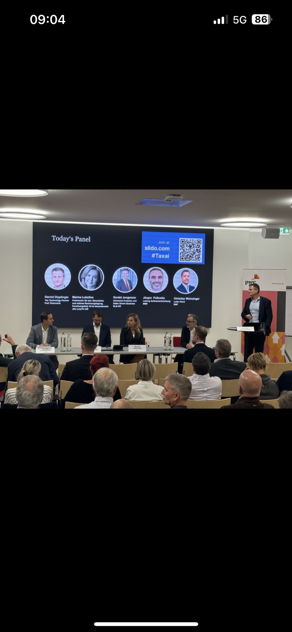 Blick von hinten auf das Publikum. Vorne sitzen mehrere Vortragende an einem Tisch, eine Person steht am Rednerpult. Auf der Präsentationsfolie ist der Titel "Today's Panel" zu sehen.