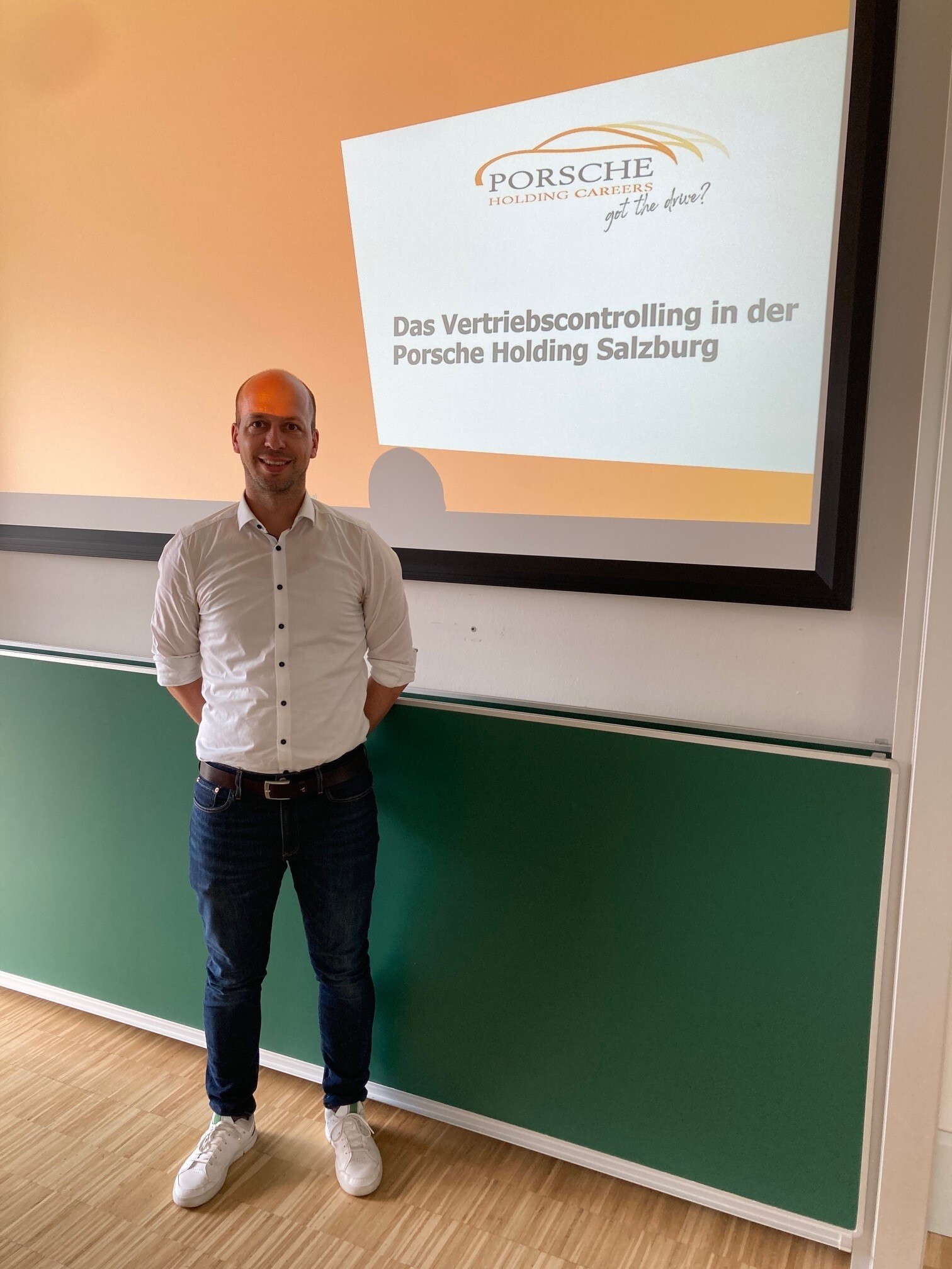 Paul Schiel von der Porsche Holding steht vor einer Leinwand. Darauf sind das Unternehmenslogo und der Titel des Vortrags „Vertriebscontrolling in der Porsche Holding Salzburg“ zu sehen.