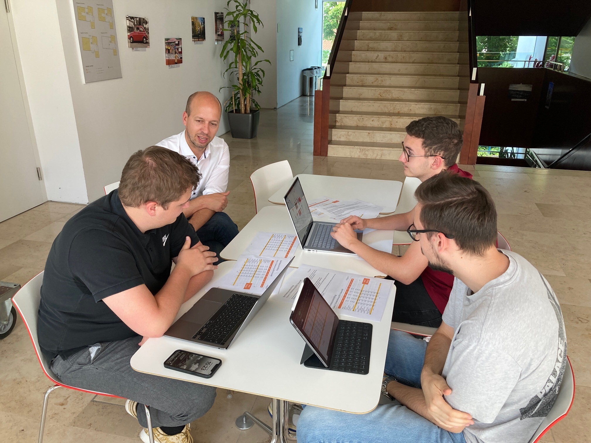 Eine Studentengruppe arbeitet gemeinsam an einem Tisch. Mehrere Personen schauen auf Unterlagen oder Laptops.