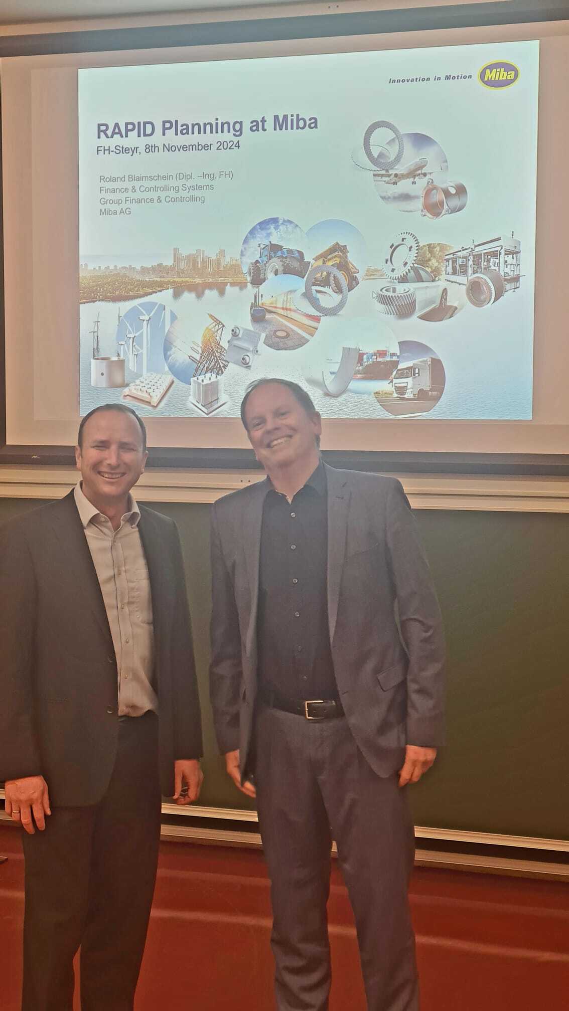 zwei Vortragende stehen vor der Tafel. Im Hintergrund ist die Titelfolie ihrer Präsentation mit der Schrift "RAPID Planning at MIBA" zu sehen.