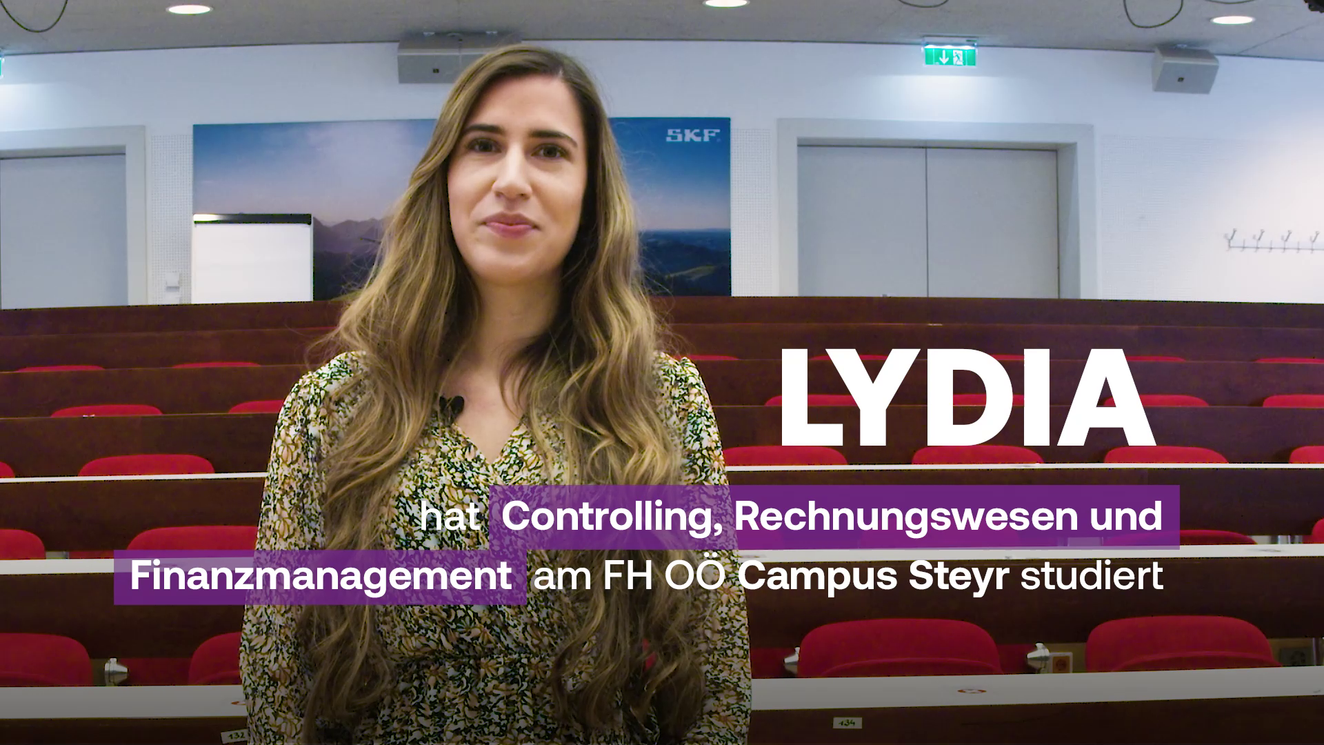 Eine Studentin steht in einem Hörsaal. Rechts neben ihr steht in weißer Schrift: "Lydia" und darunter: "hat Controlling, Rechnungswesen und Finanzmanagement am FH OÖ Campus Steyr studiert."