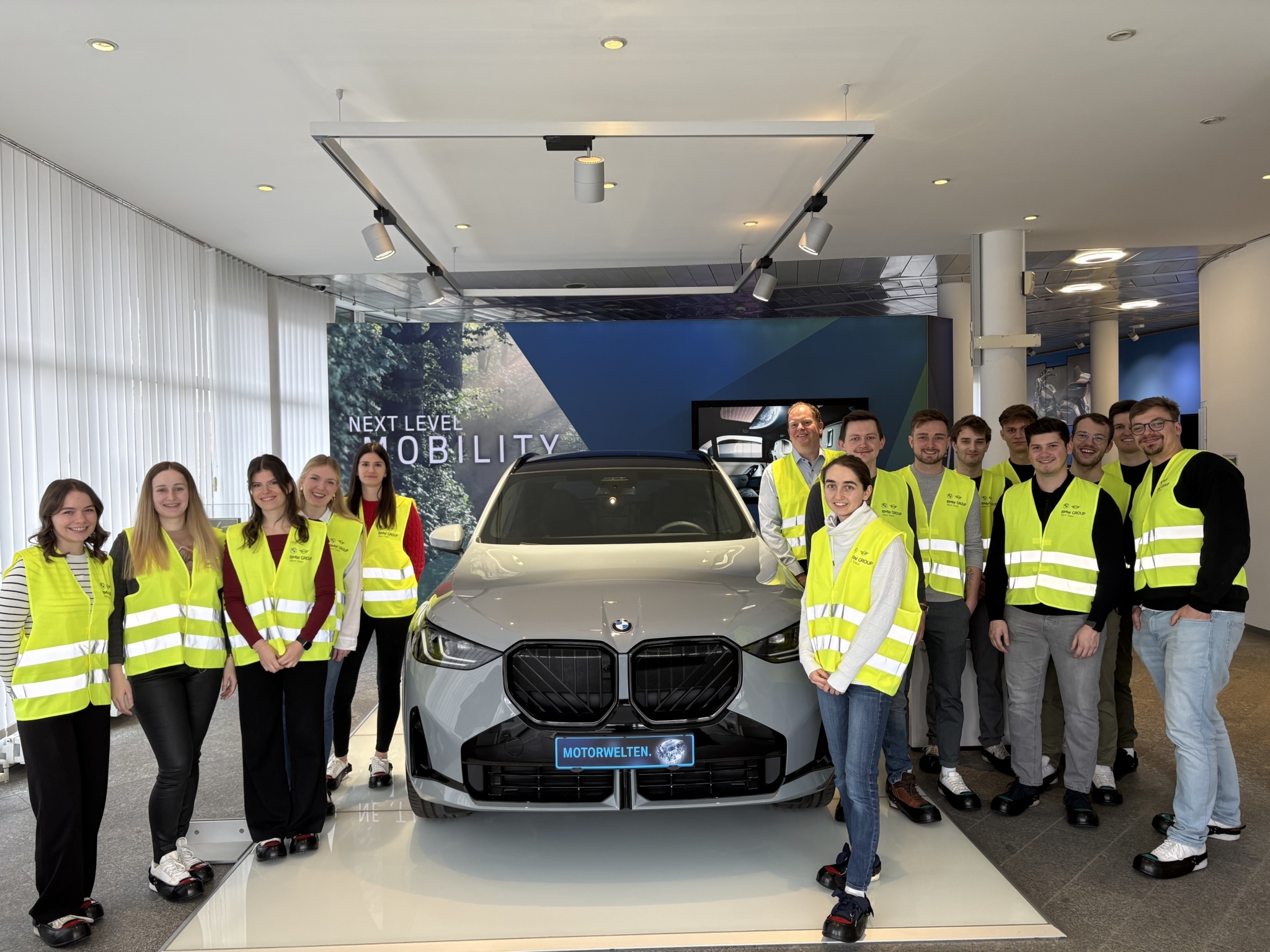 Eine Studentengruppe mit Warnwesten posiert für ein Gruppenfoto im Werk der BMW Group in Steyr. In der Mitte der Gruppe steht ein Ausstellungsauto von BMW.