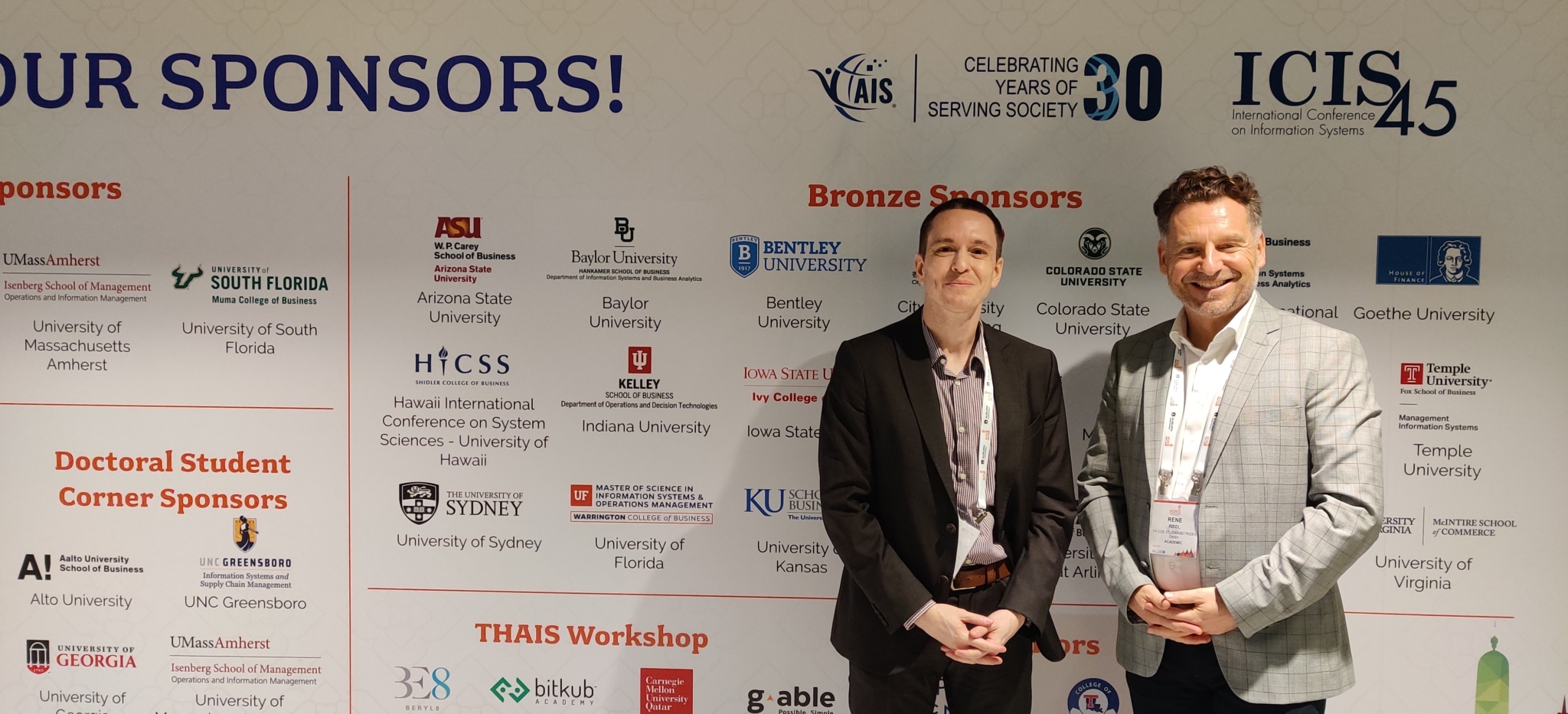 Zwei Personen stehen vor einer Wand, auf der die Logos aller Sponsoren der International Conference on Information Systems (ICIS) in Bangkok abgebildet sind.