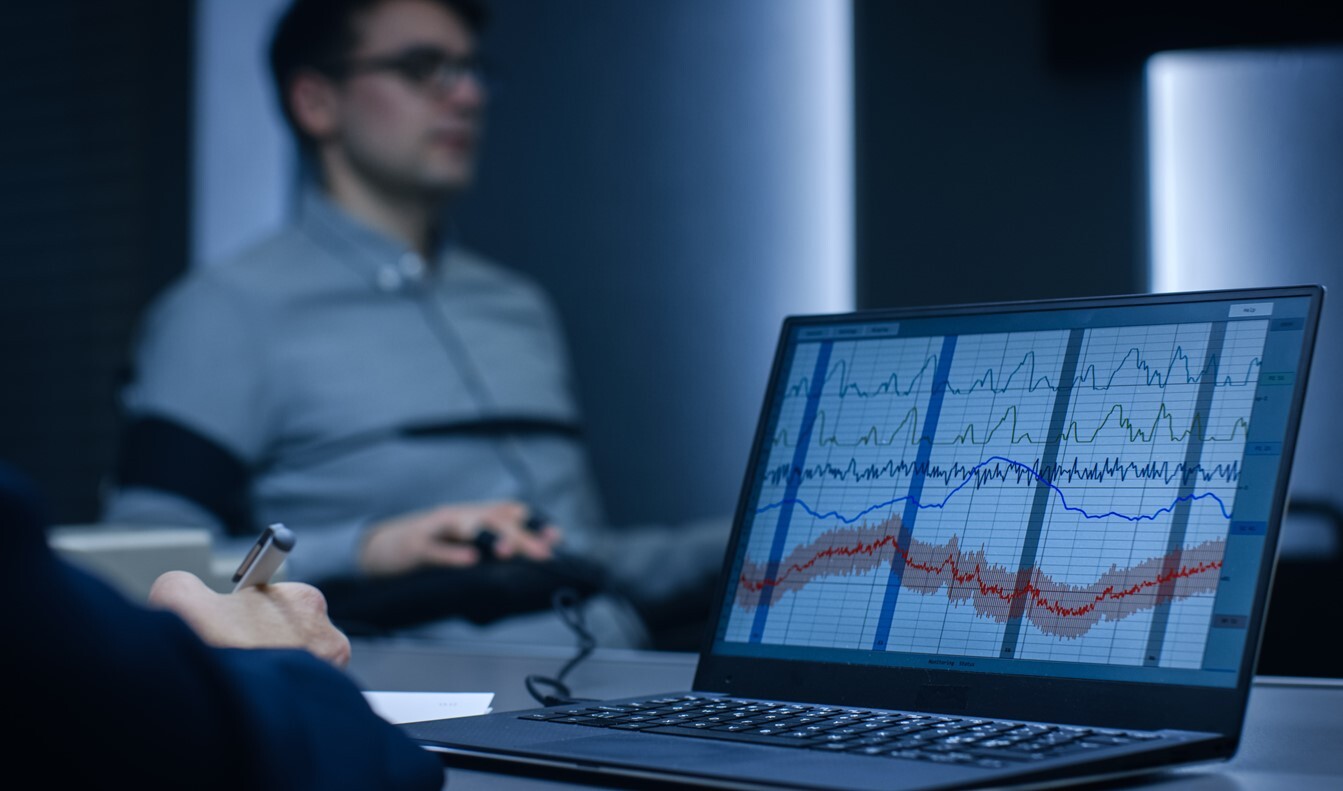 Ein Laptop zeigt Diagramme und Kurven, die Messwerte der im Hintergrund sitzenden Person darstellen. Die Person trägt eine Brille und ist mit Sensoren oder Kabeln verbunden.