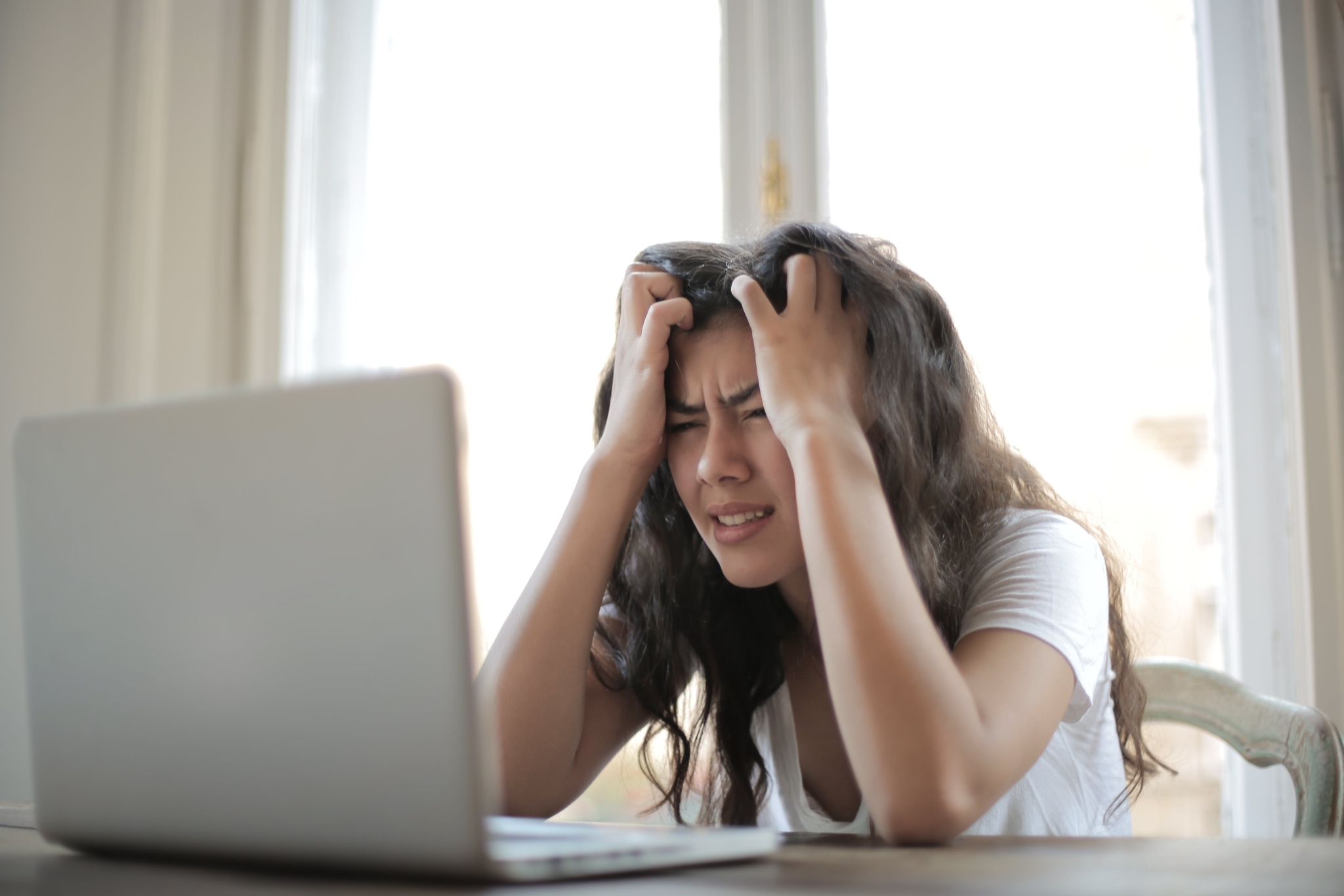 Eine junge Frau sitzt vor einem Laptop, stützt den Kopf mit den Händen ab und blickt verzweifelt auf den Bildschirm.