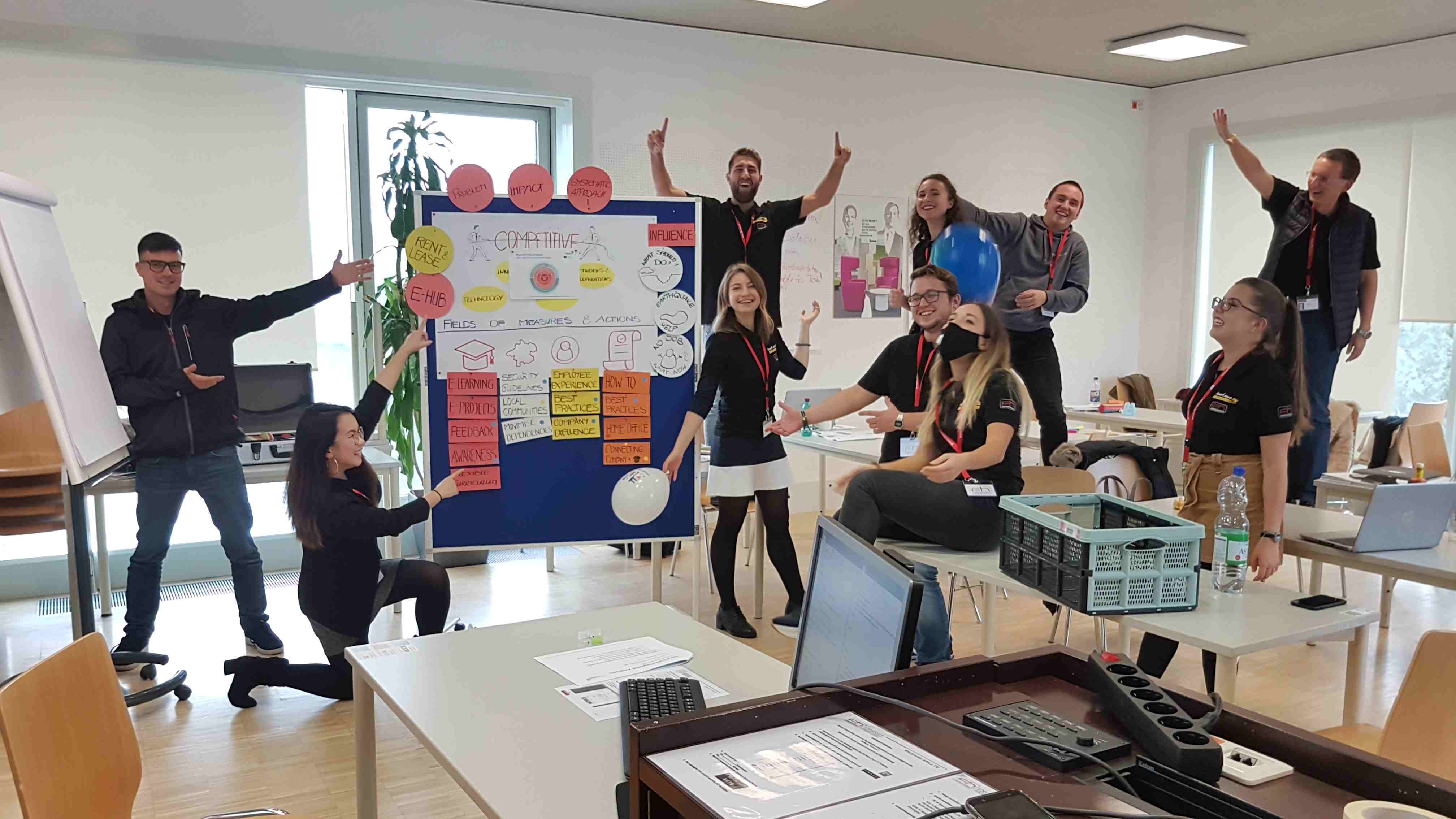 Group of students in a seminar room full of energy and joy. A pinboard with diagrams and notes can be seen in the background. The room is furnished with tables and chairs.