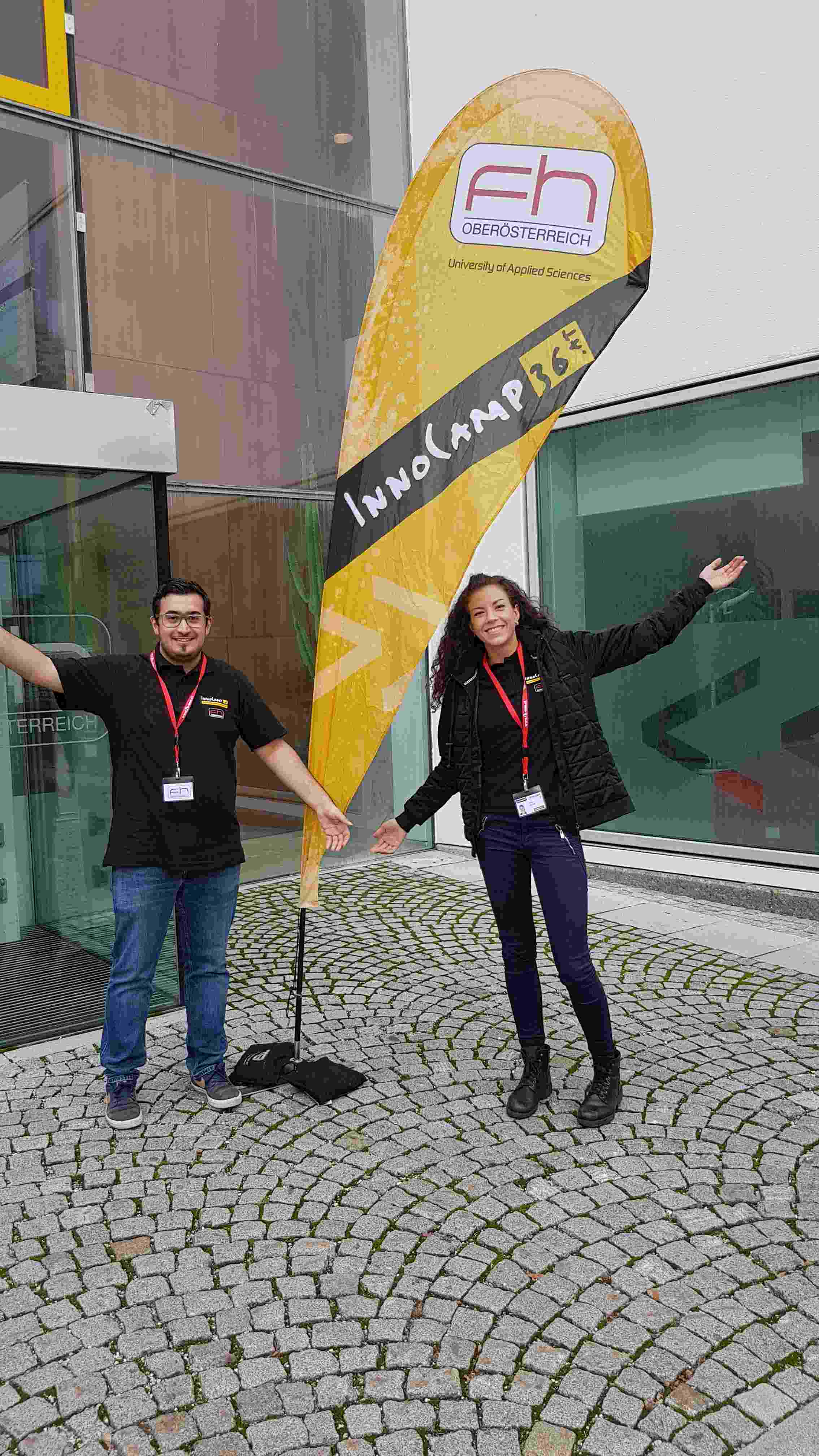 Two people stand in front of the FH2 building with their hands outstretched from a display of the FH Upper Austria Innocamp36