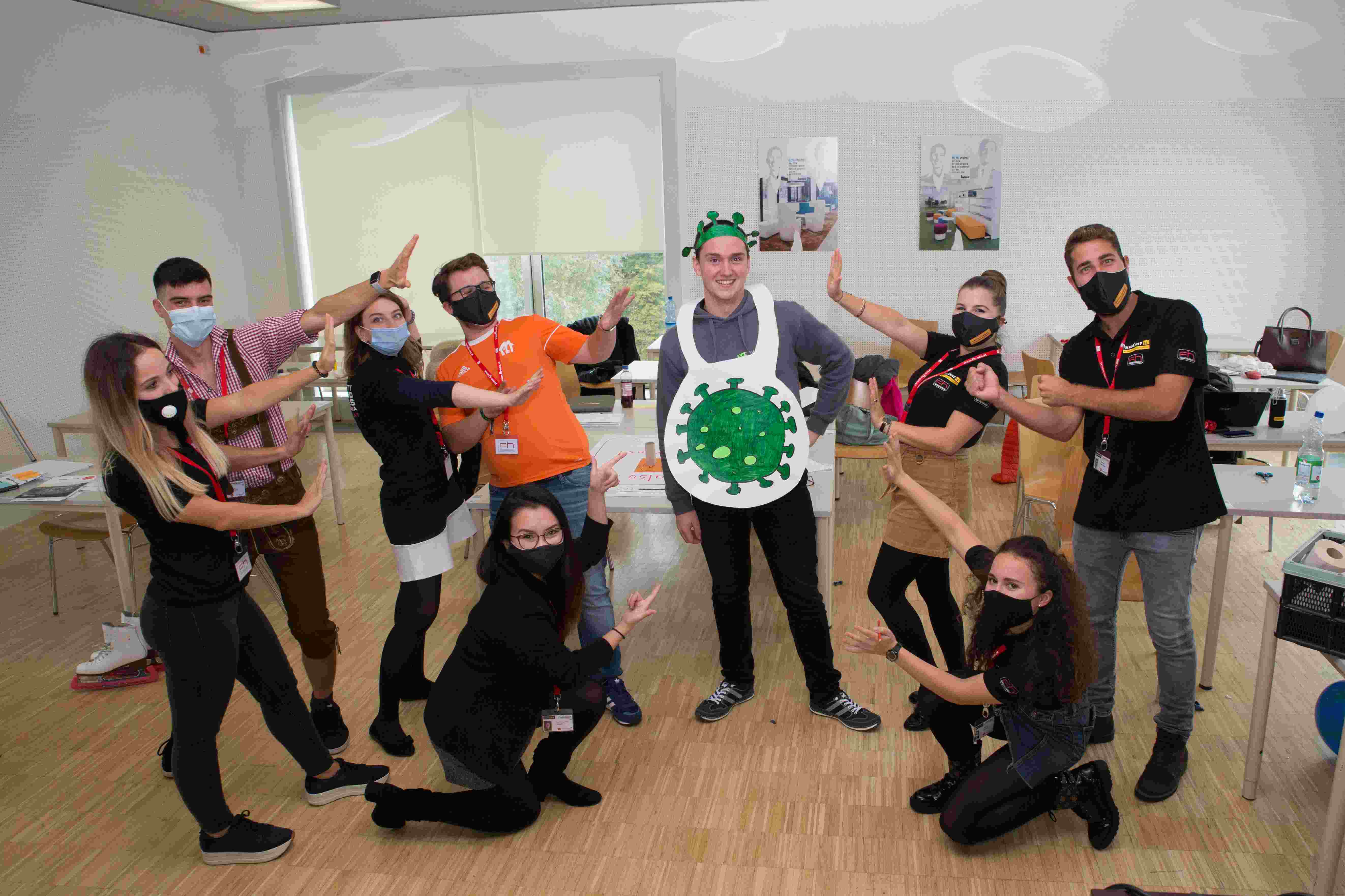 Group of students posing for a photo in the seminar room. The student in the centre is wearing a costume that resembles a virus. The other students are wearing masks.