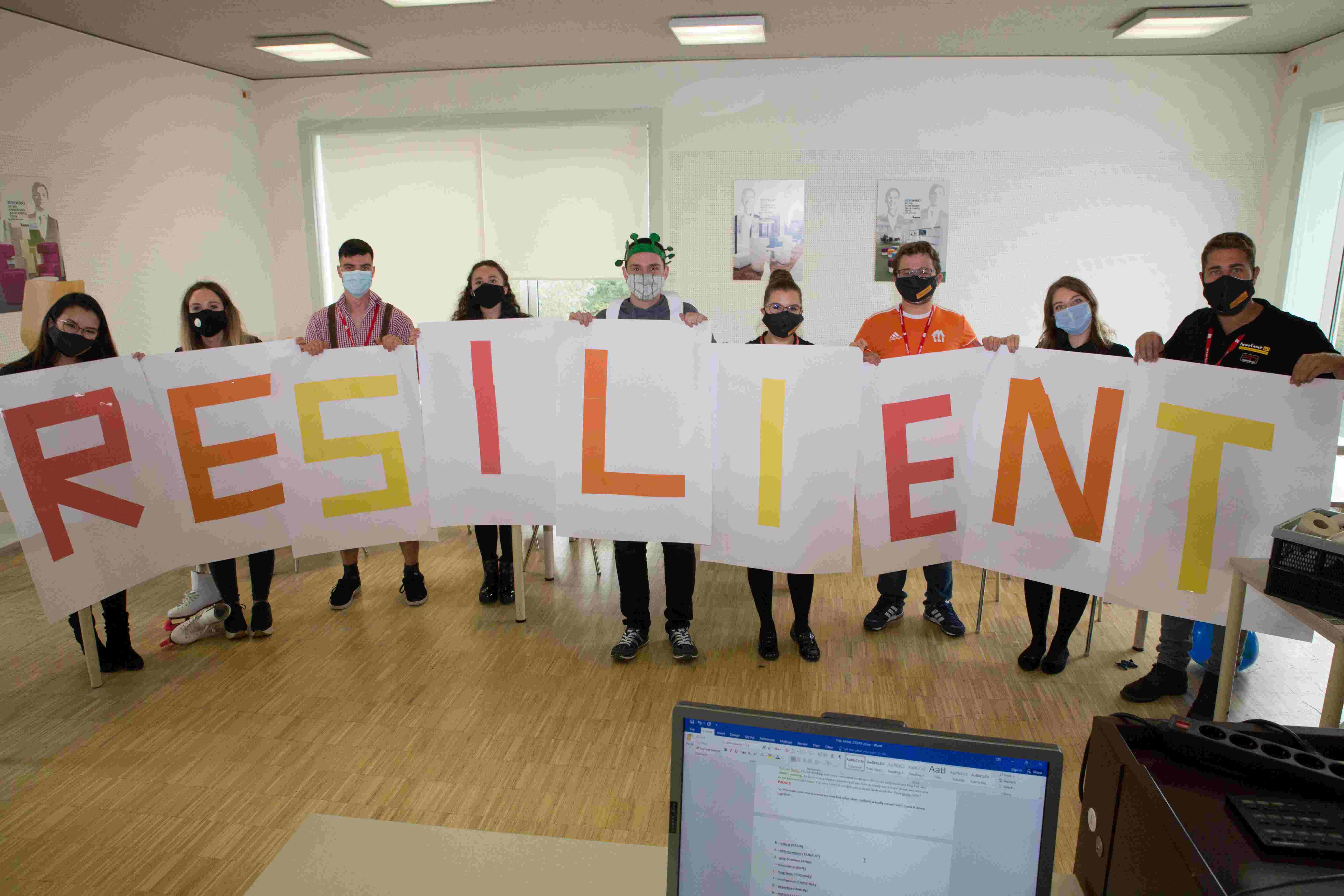 Nine people each hold a poster with a letter on it. Together, the letters form the word “Resilient”.