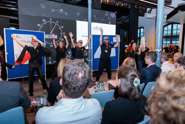 Studentengruppe, die auf einer Bühne performt, während ein Publikum zuschaut. Die Performer haben die Arme erhoben und präsentieren eine Show.
