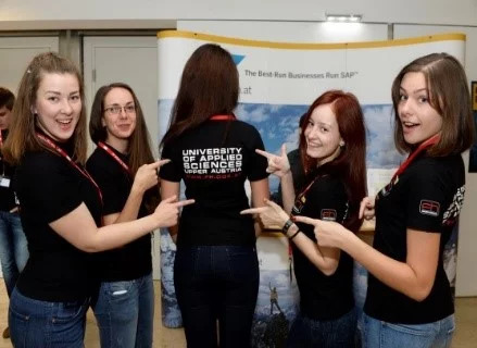 Eine Studentin steht mit dem Rücken zur Kamera und trägt ein T-Shirt mit dem Logo der Fachhochschule. Um sie herum stehen weitere Studierende, die in die Kamera lächeln und auf das Logo auf dem T-Shirt zeigen.