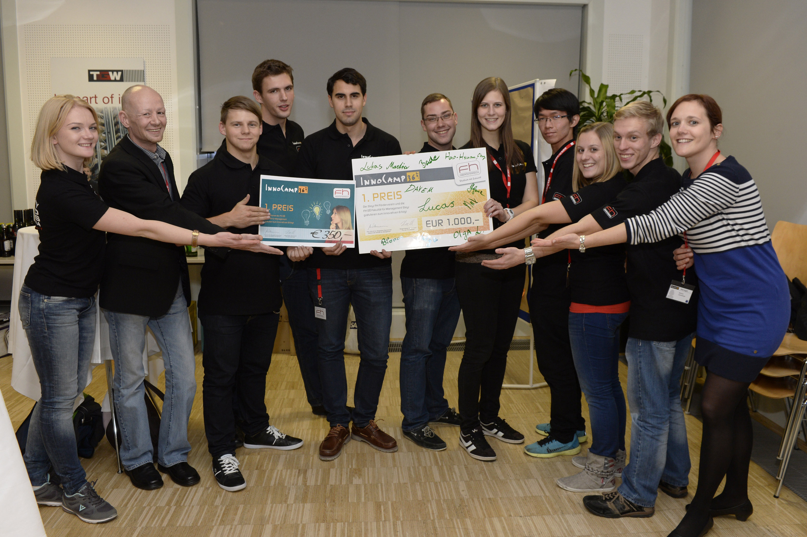 A group, most of them wearing black T-shirts with orange logos, stand in a row smiling and holding awards and cheques up to the camera.