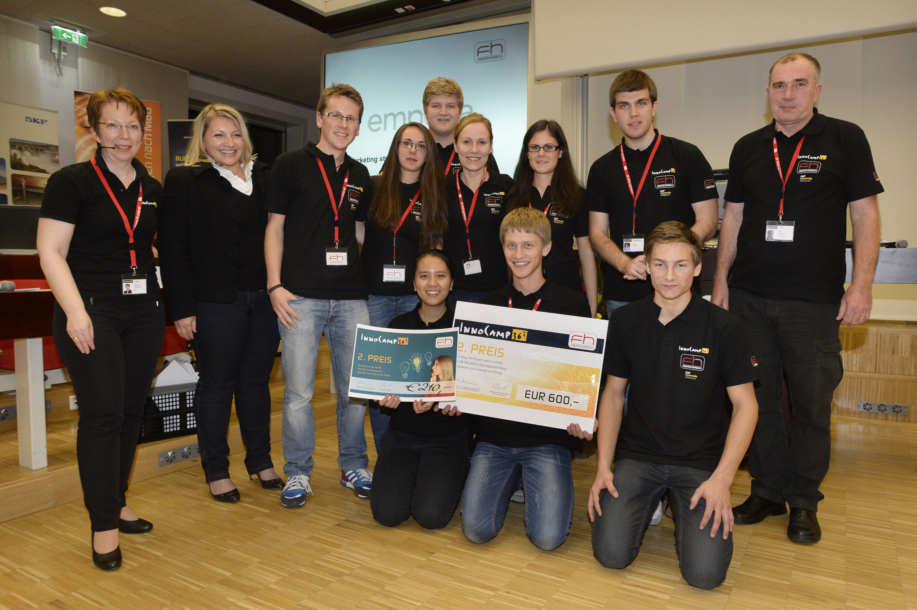 A group of people, most of them wearing black T-shirts with orange logos, stand in a row smiling and holding awards and cheques up to the camera.