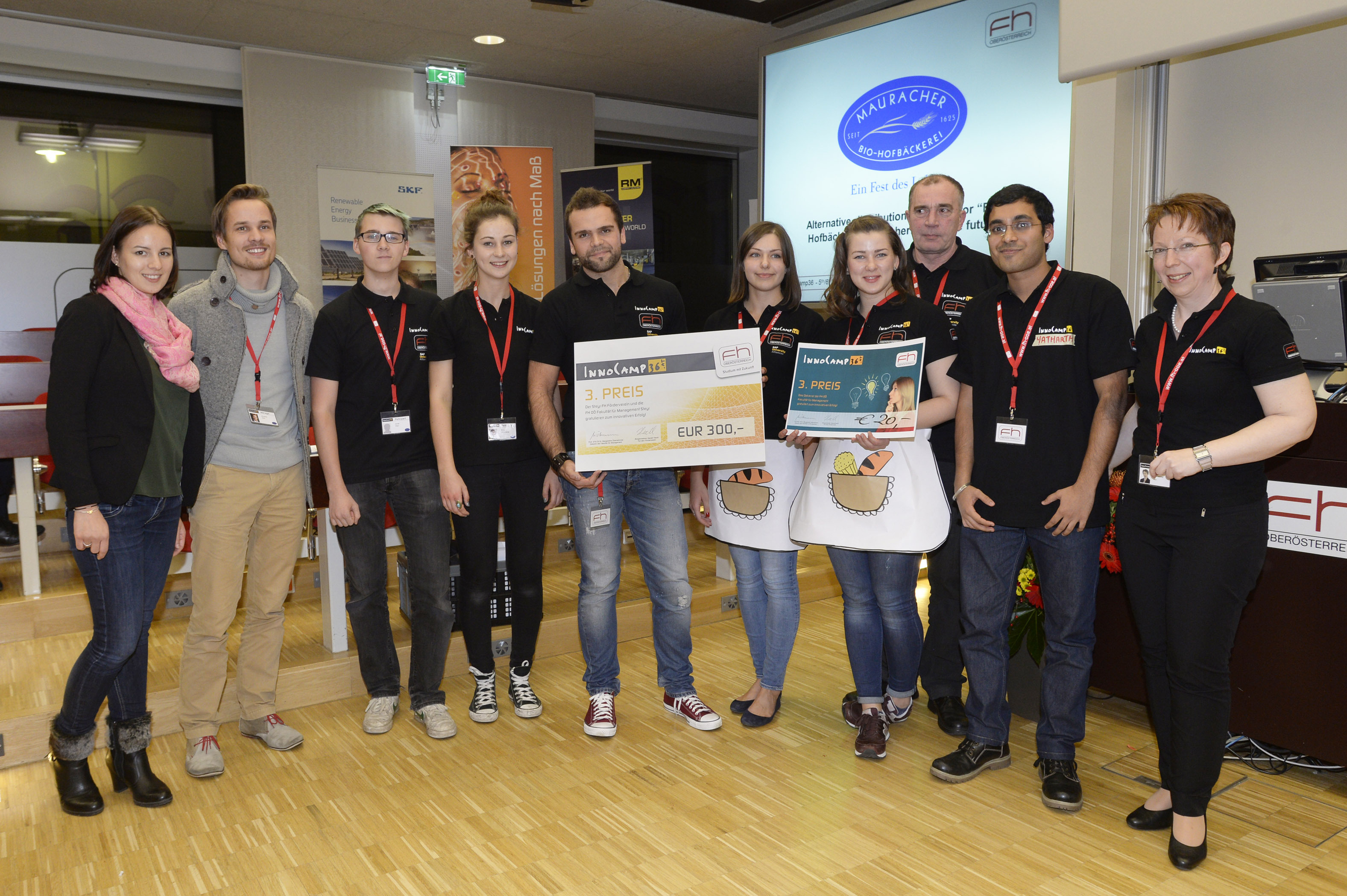 A group of eleven people, most of them wearing black T-shirts with orange logos, stand in a row smiling and holding awards and cheques up to the camera.