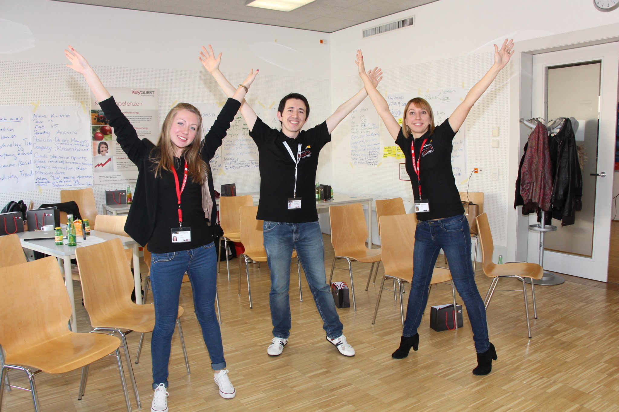 Two female and one male student stand in a seminar room and raise their arms in the air as if they are cheering.