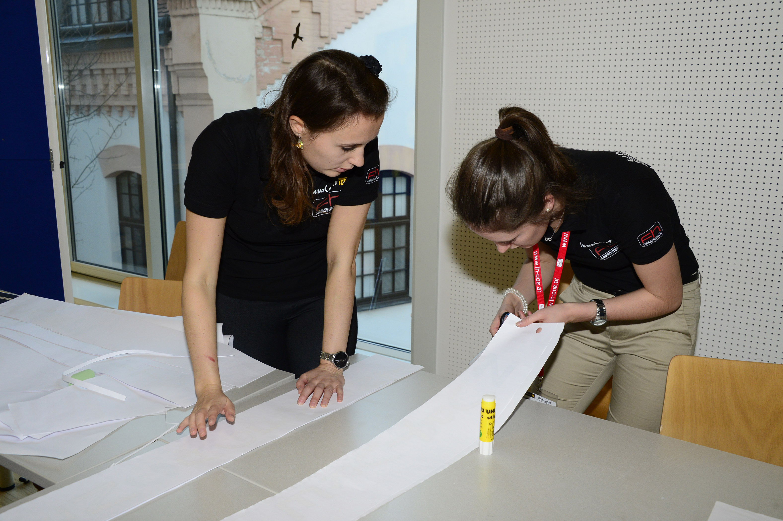 Two young women work at a table cutting white paper