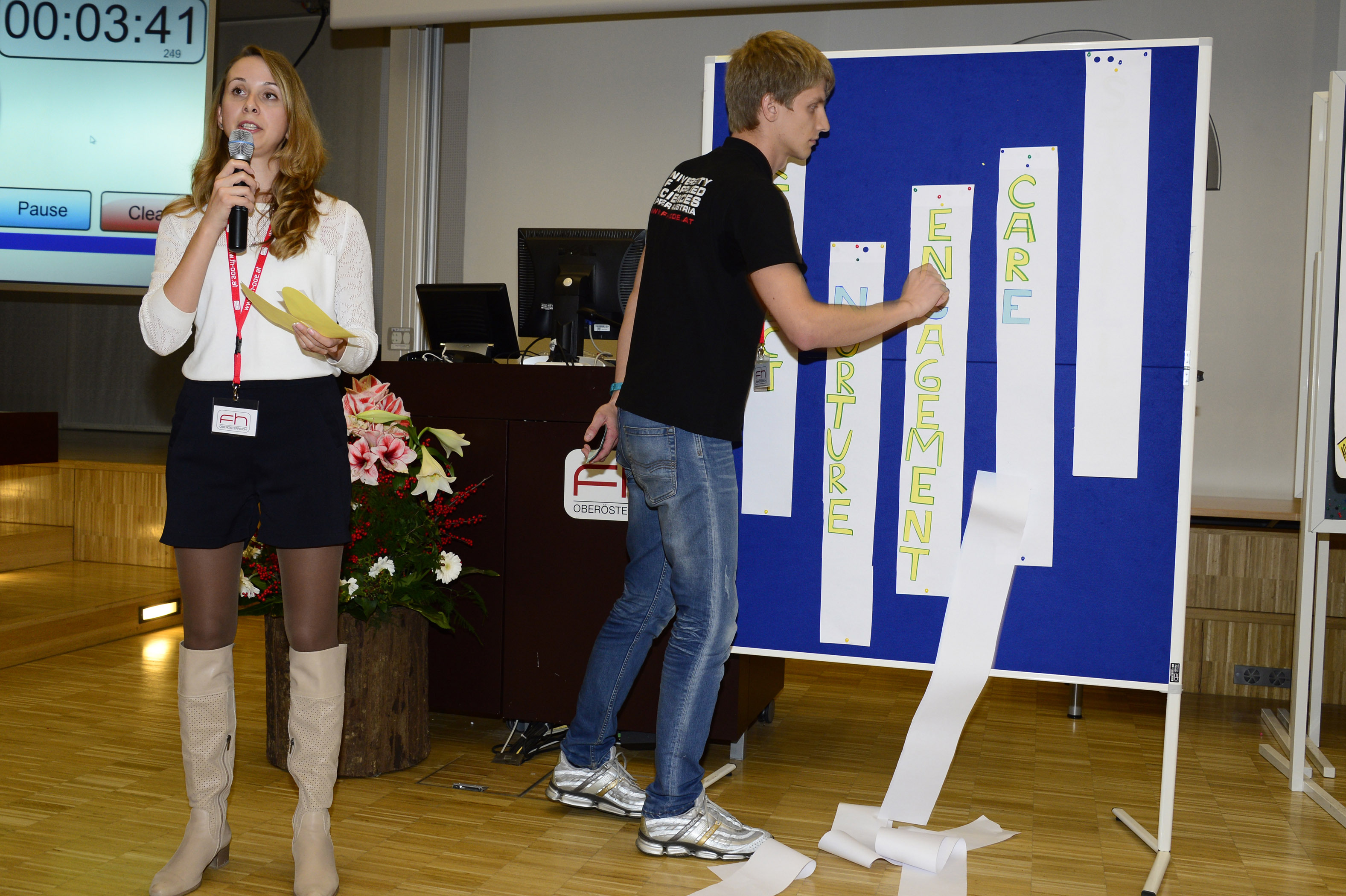 A young female student holds a microphone in her hand as she presents. Next to her, a young man stands in front of a blue pinboard and attaches long white strips of paper with words such as ‘CARE’, “ENGAGEMENT” and ‘FUTURE’ written on them.