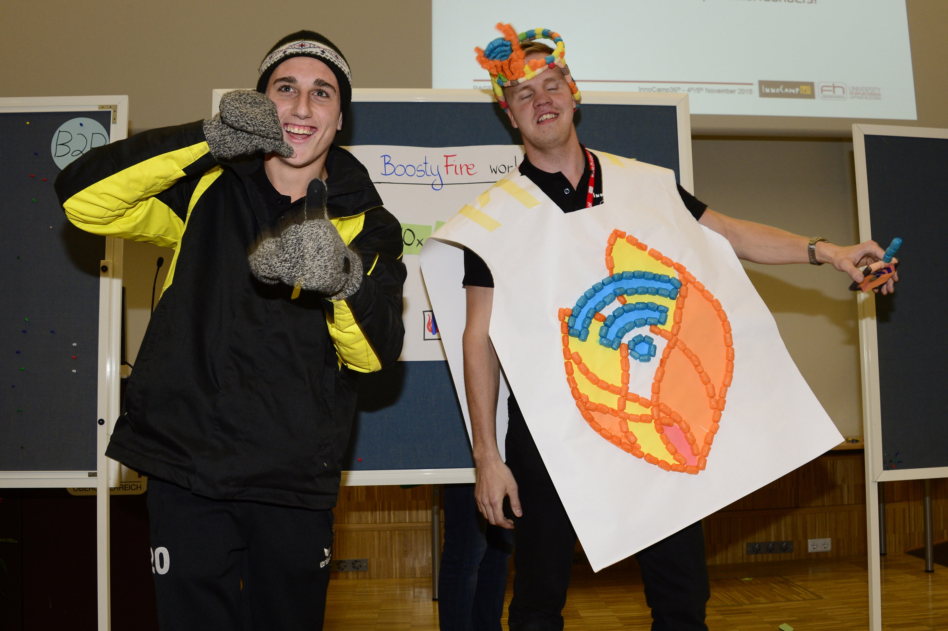 Two people in creative costumes stand next to each other. The person on the left is wearing a hat and gloves and a black and yellow jacket. The person on the right is wearing a white cape-like costume with a large, colourful symbol that combines flames and a Wi-Fi signal.