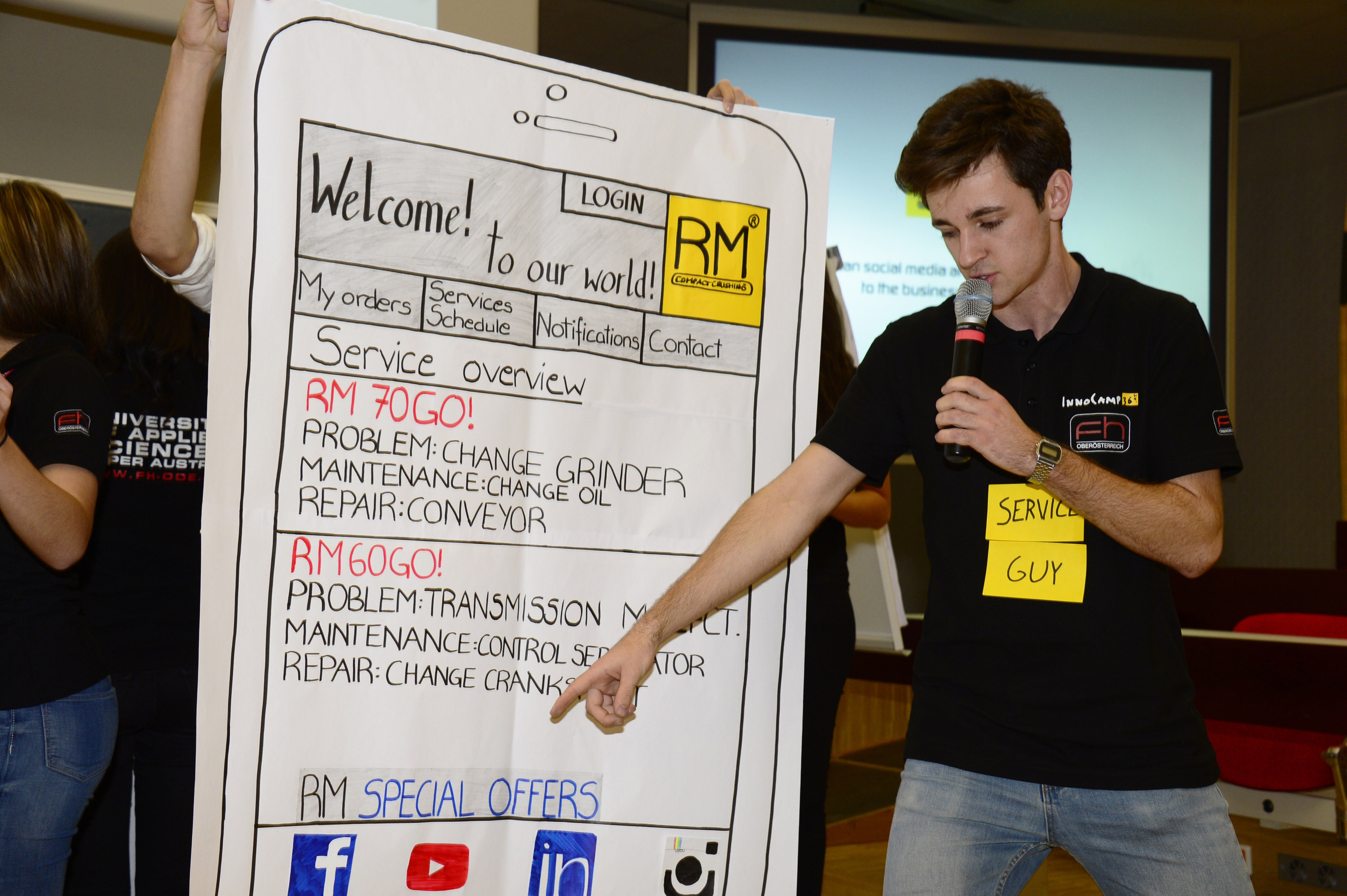 A young student with a microphone in his hand points to a large poster designed like the screen of a smartphone. The poster shows headlines such as "Welcome! to our world!‘, “LOGIN”, ’Service overview" and lists technical problems and repairs. The student is wearing a black T-shirt and jeans, and a yellow sticker with the words ‘SERVICE GUY’ is stuck to his T-shirt.