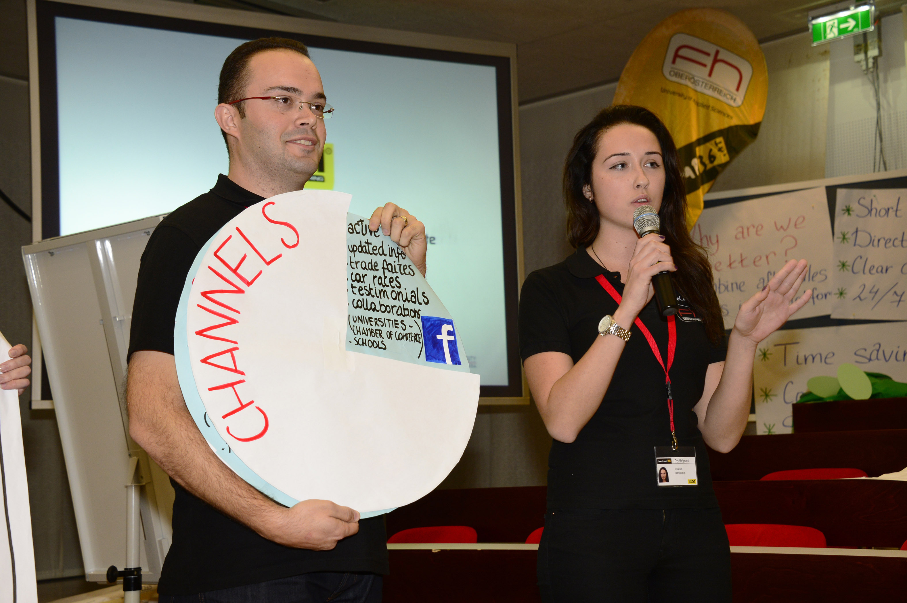 A male and female student are standing in front of a screen. The man is holding a large, circular sign with the word ‘CHANNELS’ on it, on which various sources of information and a Facebook logo are depicted. The woman is holding a microphone. Both are wearing black T-shirts. A projection screen and other boards can be seen in the background.