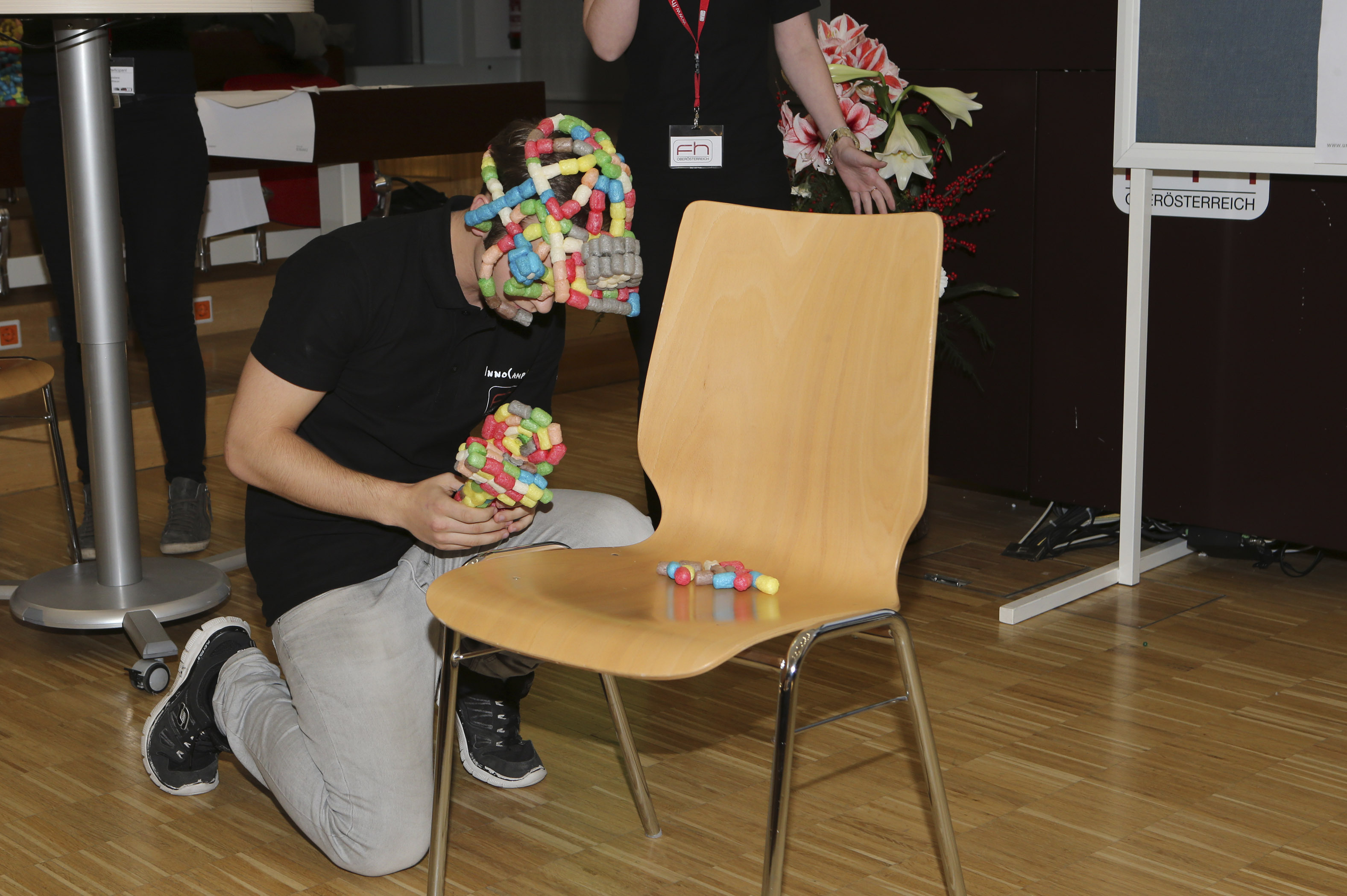 A student on his knees on the floor in front of a wooden chair. On his head is a colourful, curious headgear. He is holding a similar object in his hands. There are also some of these colourful elements on the seat of the chair. Other people and a white whiteboard can be seen in the background.
