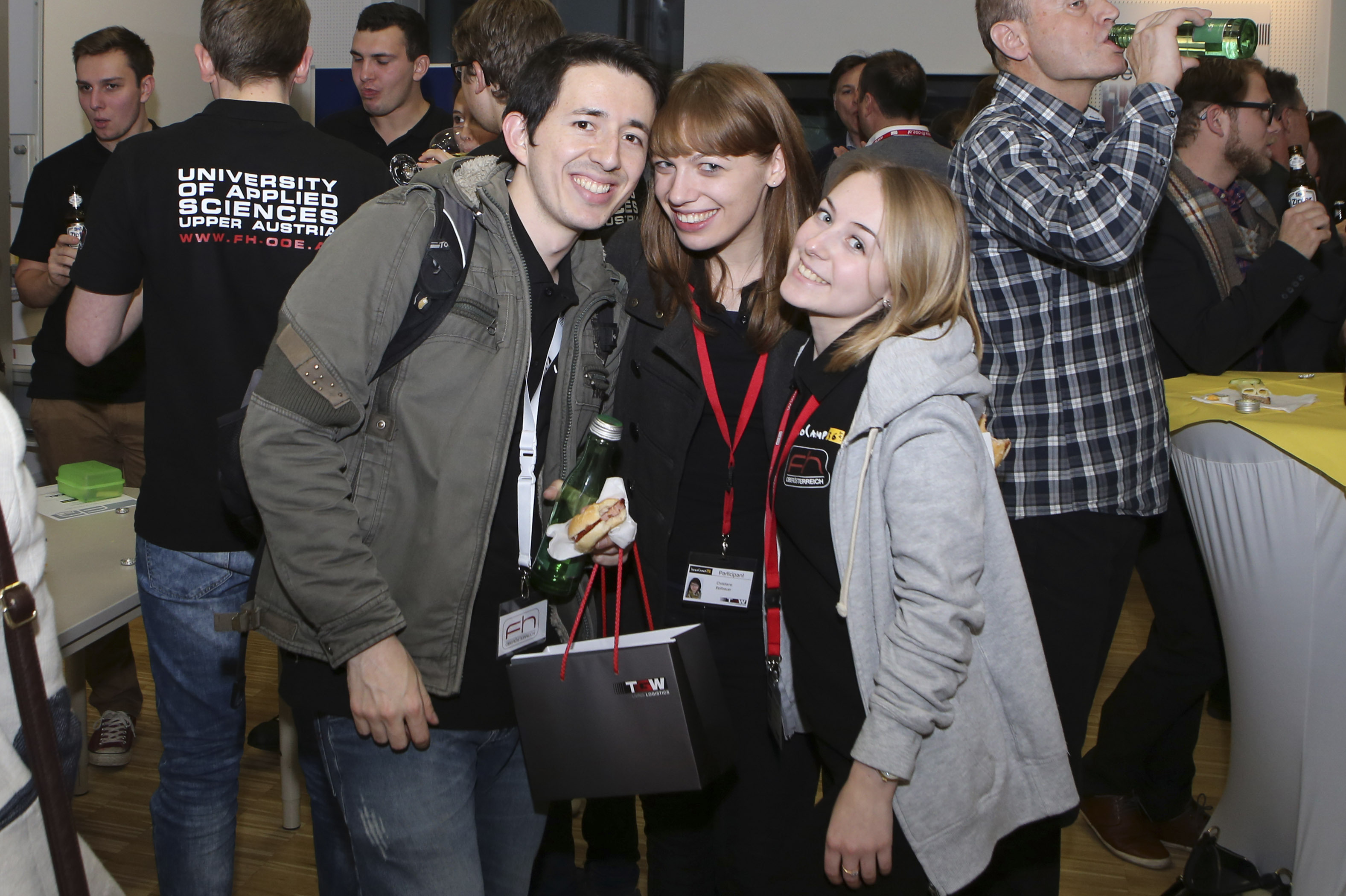Three students are smiling and posing for a photo, surrounded by other people in the background.